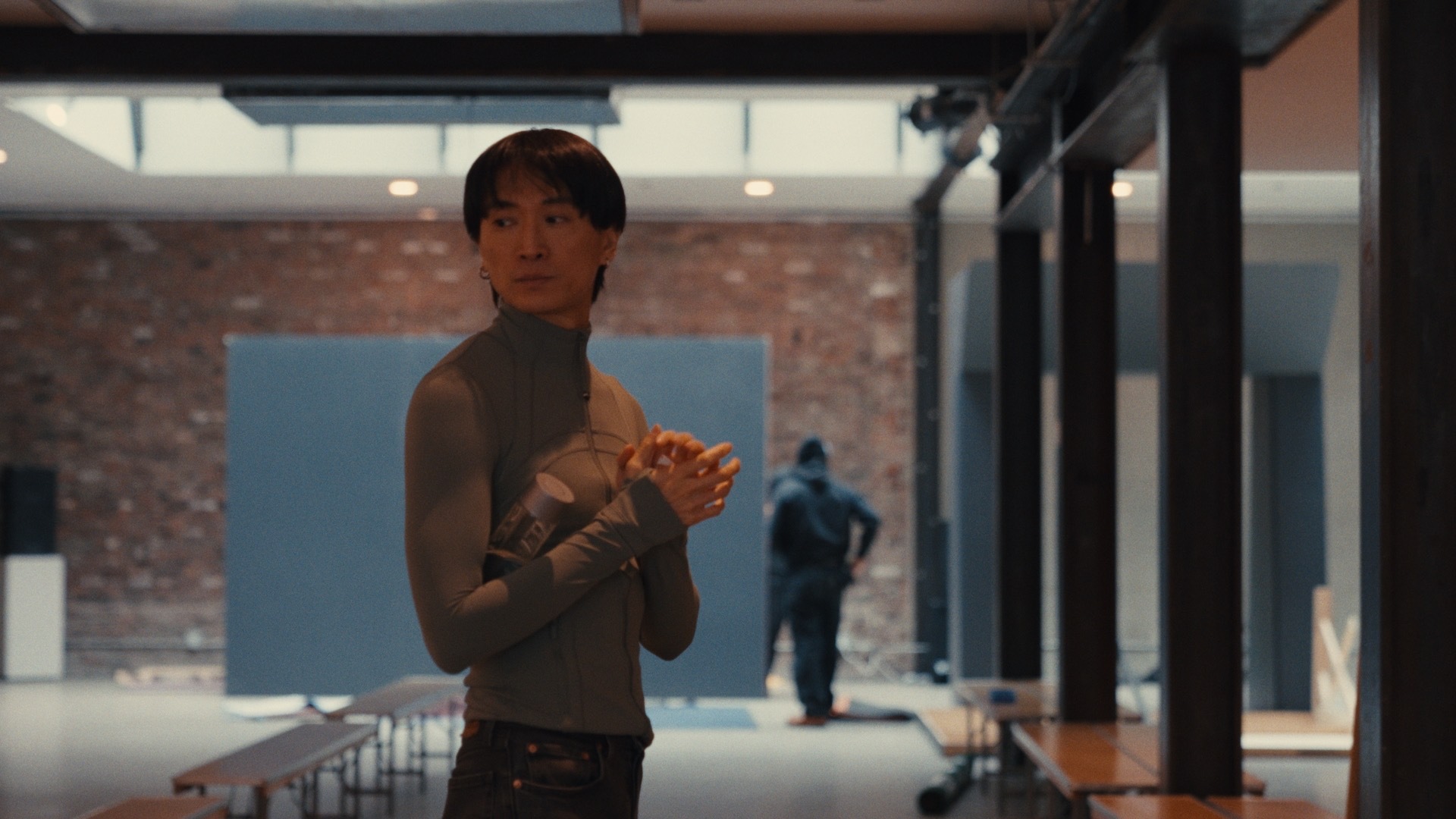 Person standing in a large industrial-style room with hands clasped, holding a water bottle under one arm, while another person appears blurred in the background