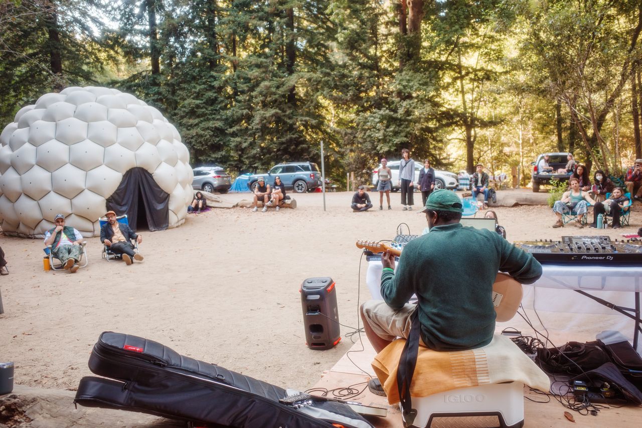 Musician plays guitar to a small audience in a forest clearing beside a white dome tent