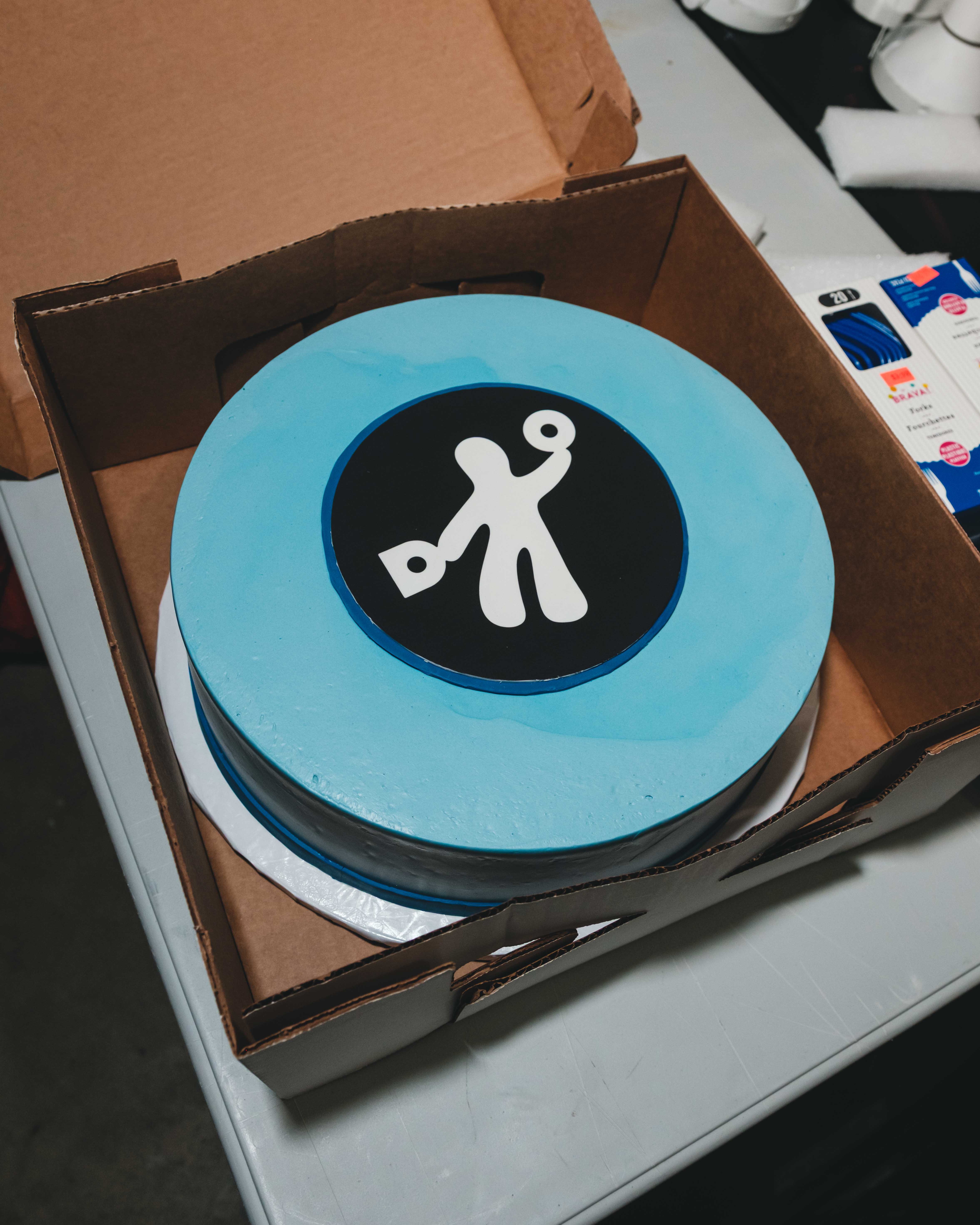 Blue round cake in an open cardboard bakery box, decorated with a black circle and white stick-figure logo on top