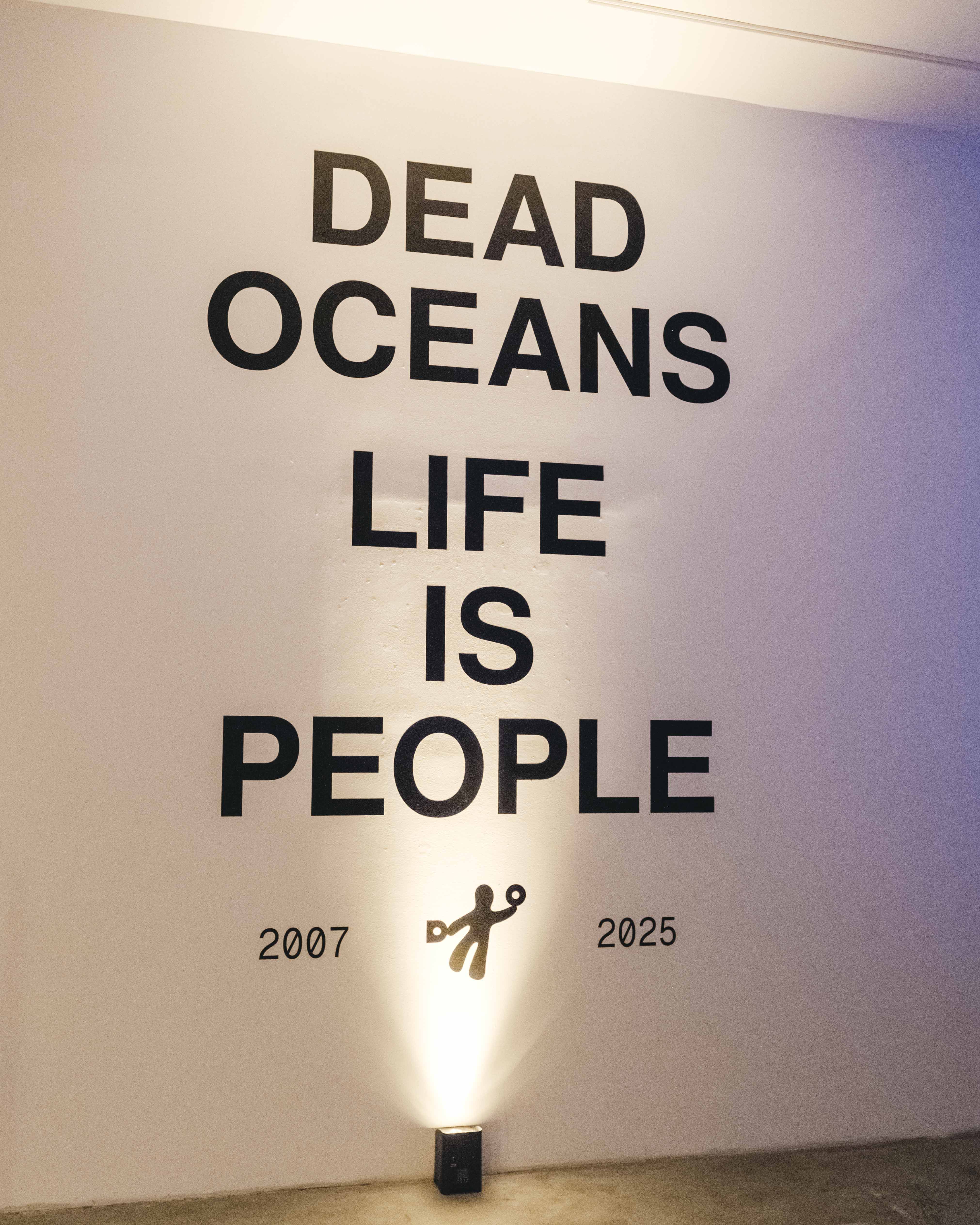 Wall installation with large black text reading “DEAD OCEANS LIFE IS PEOPLE,” with “2007” and “2025” near the bottom and a small spotlight on the floor casting light upward