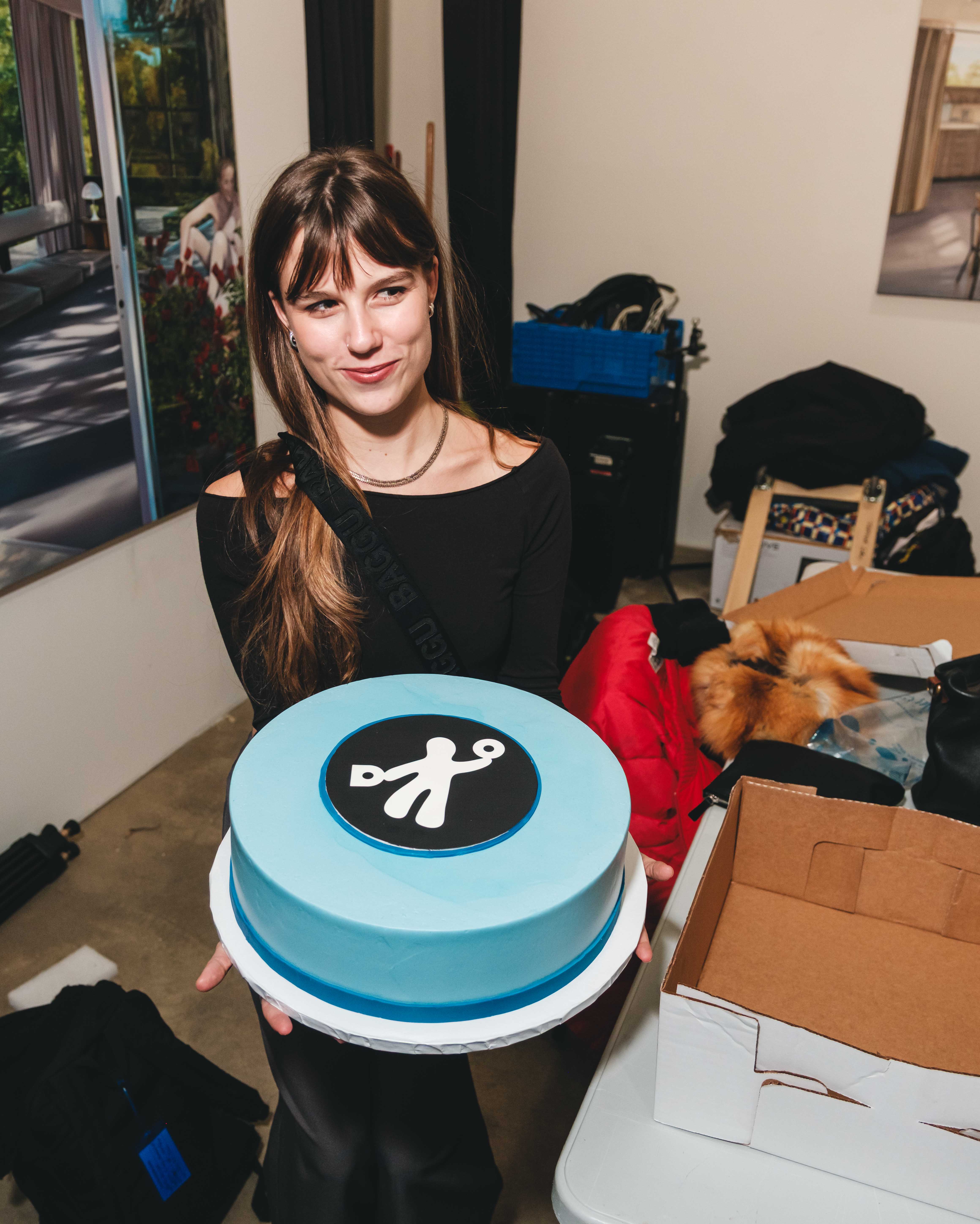 Person holding a round light blue cake with a black-and-white logo on top in an indoor event space.
