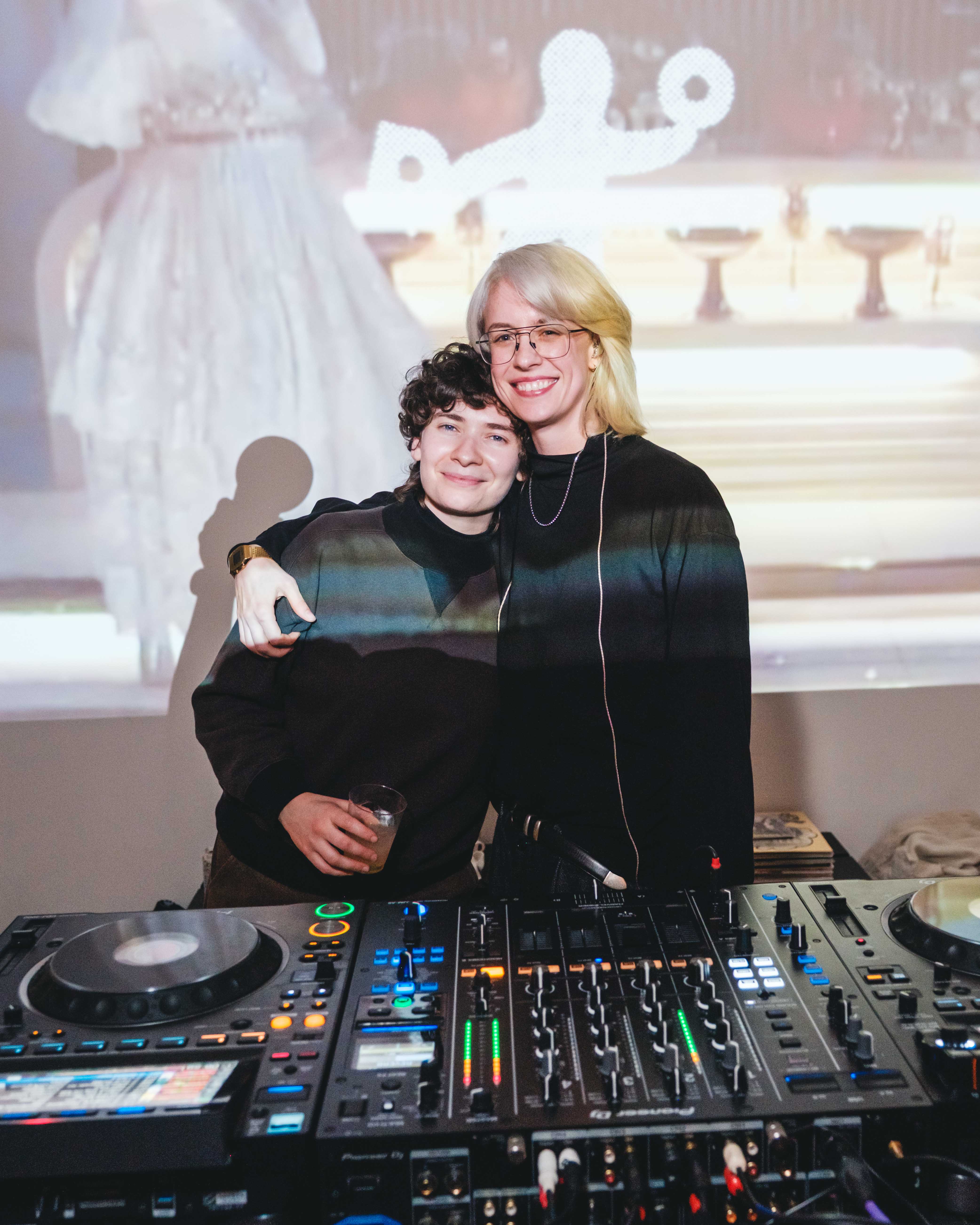 Two people stand behind a DJ booth with turntables and a mixer