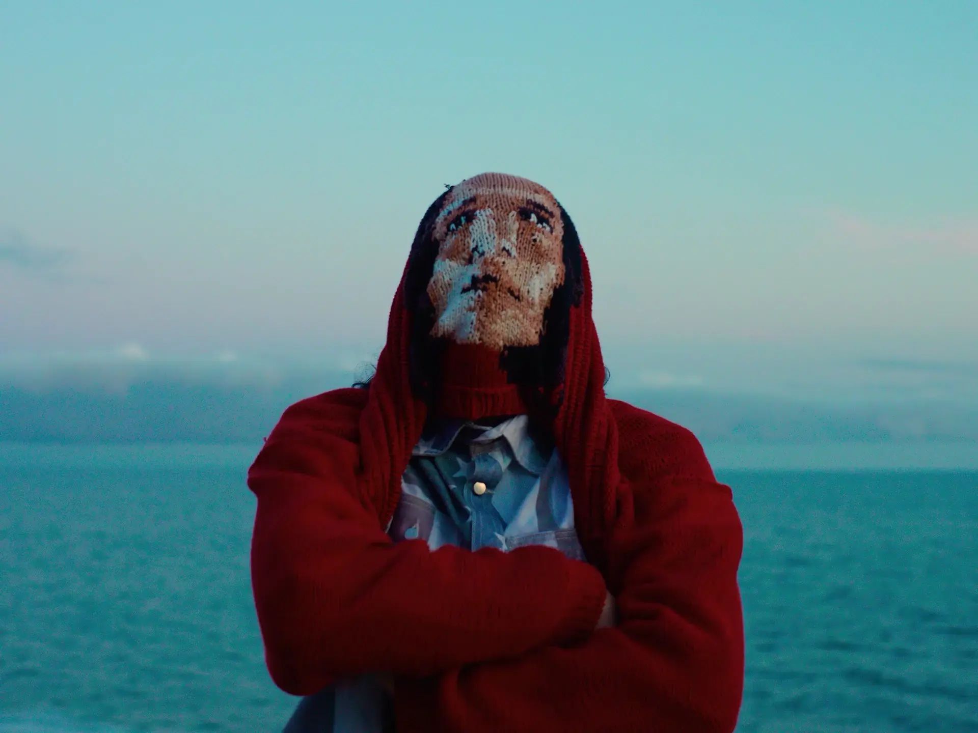Person standing by the ocean with arms crossed, wearing a red hooded sweater pulled over their face like a knit mask.