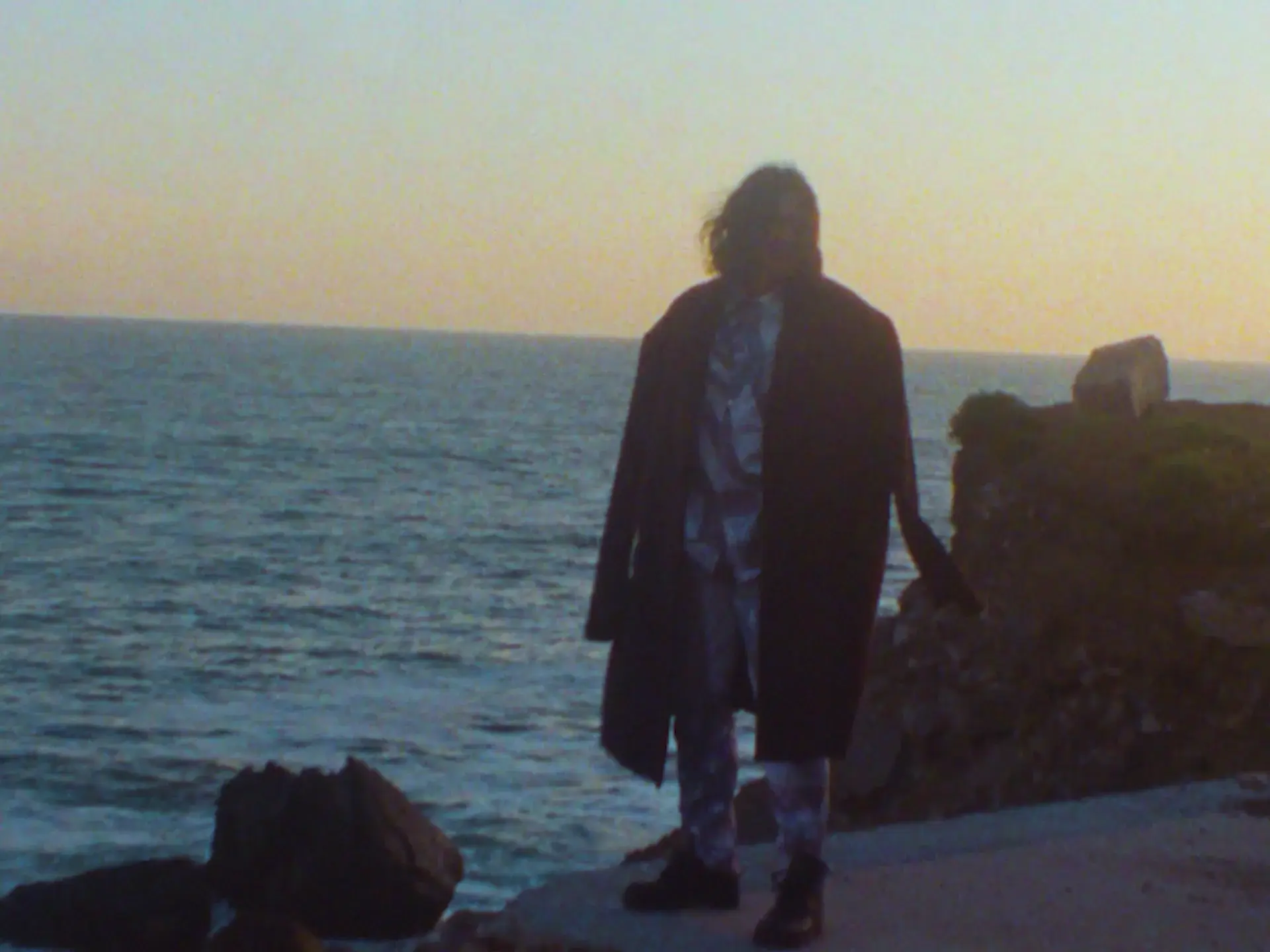 Person standing on a rocky shoreline at sunset, facing the ocean in a long dark coat and patterned clothing.