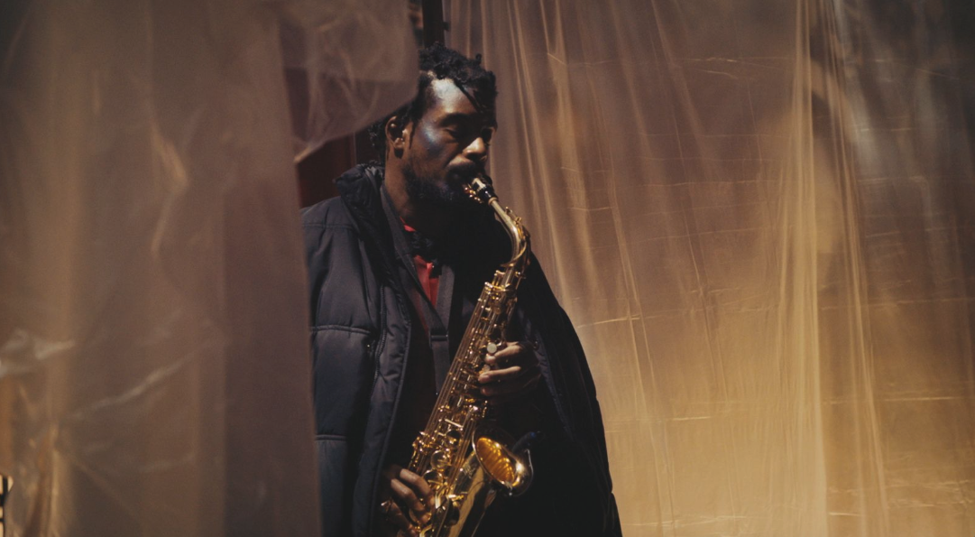 Person holding a saxophone behind sheer curtains in warm light.