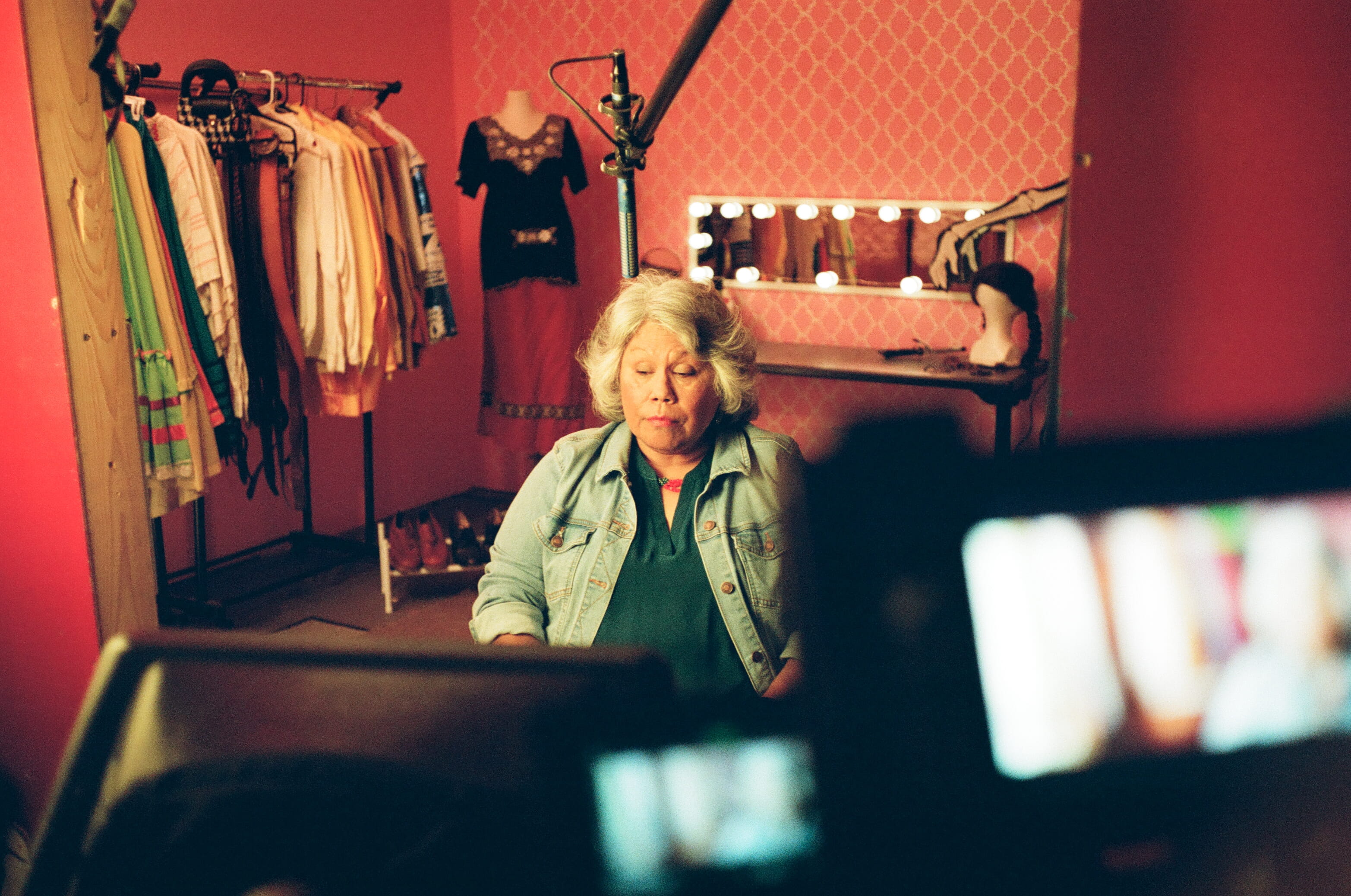 Person sitting in a dressing room with clothing racks and a lit mirror