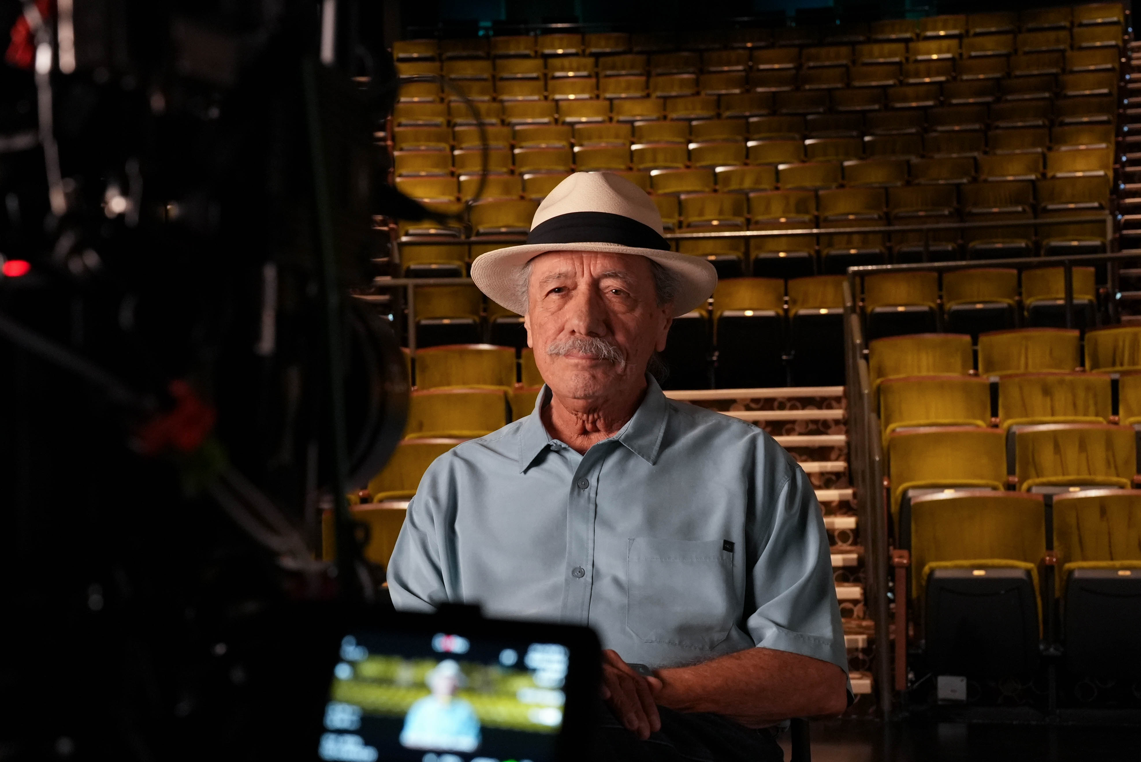 Man wearing a hat seated in a theater during a filmed interview