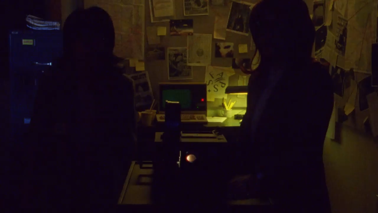Two silhouetted people stand in a dimly lit room with a desk, laptop, and wall covered in photos, notes, and clippings