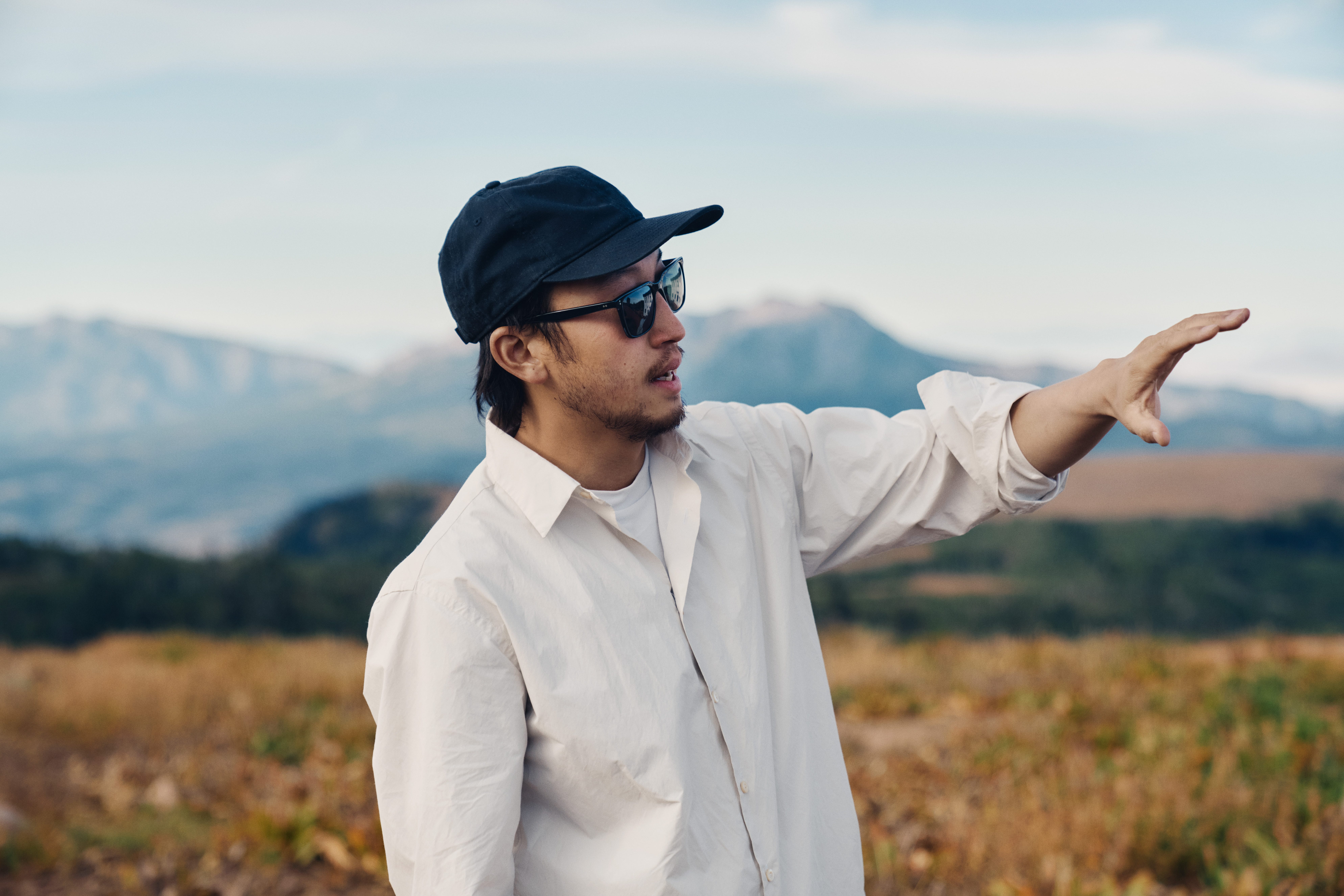 Man wearing a cap and sunglasses gesturing toward the distance in a mountainous landscape