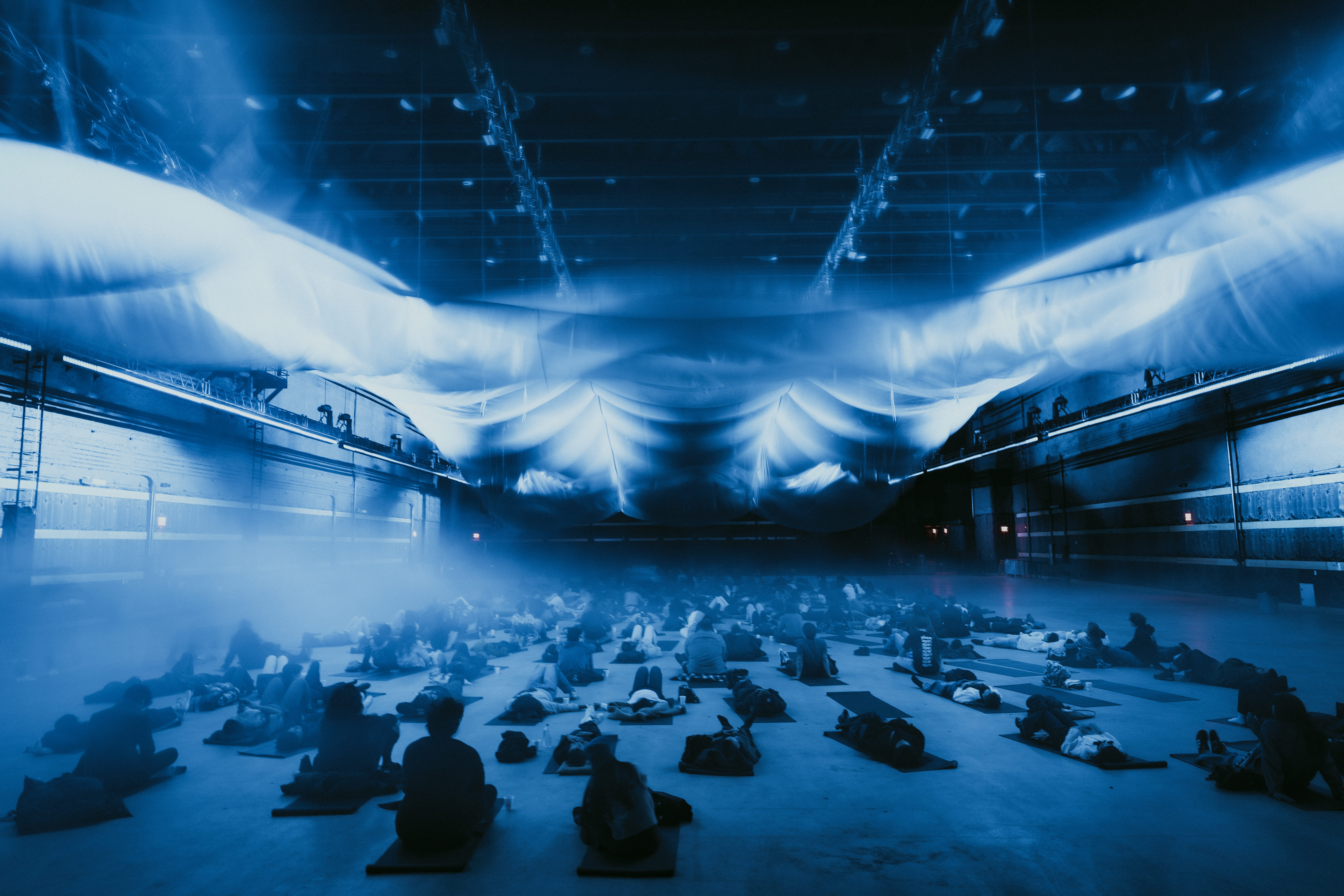 Large group seated beneath glowing blue fabric installation in a dark indoor venue