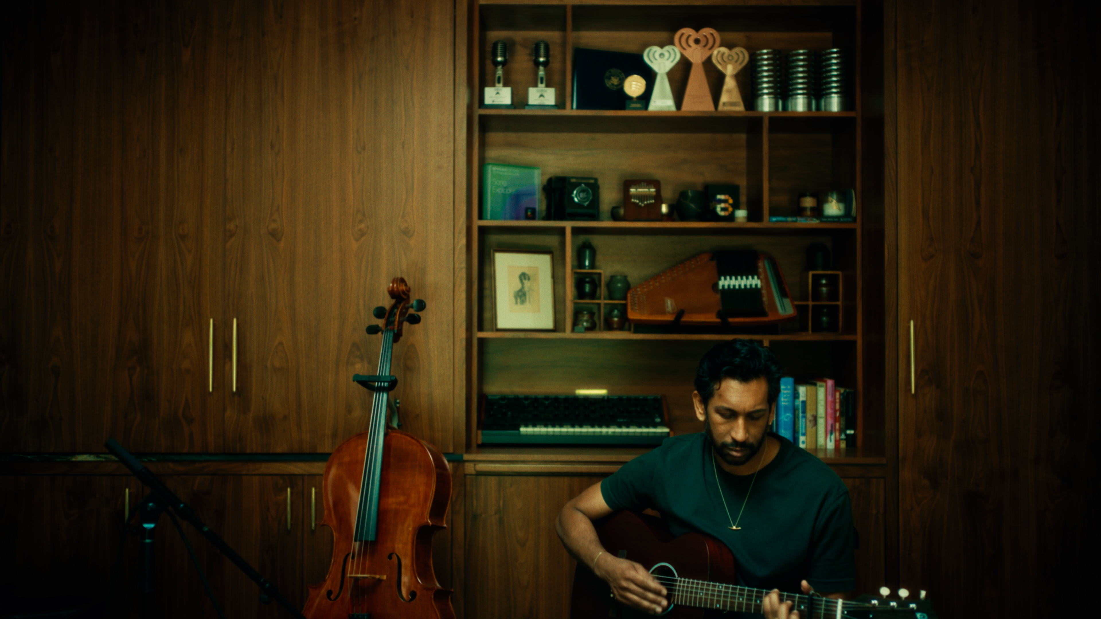 Man playing an acoustic guitar beside a cello in a wood-paneled music room