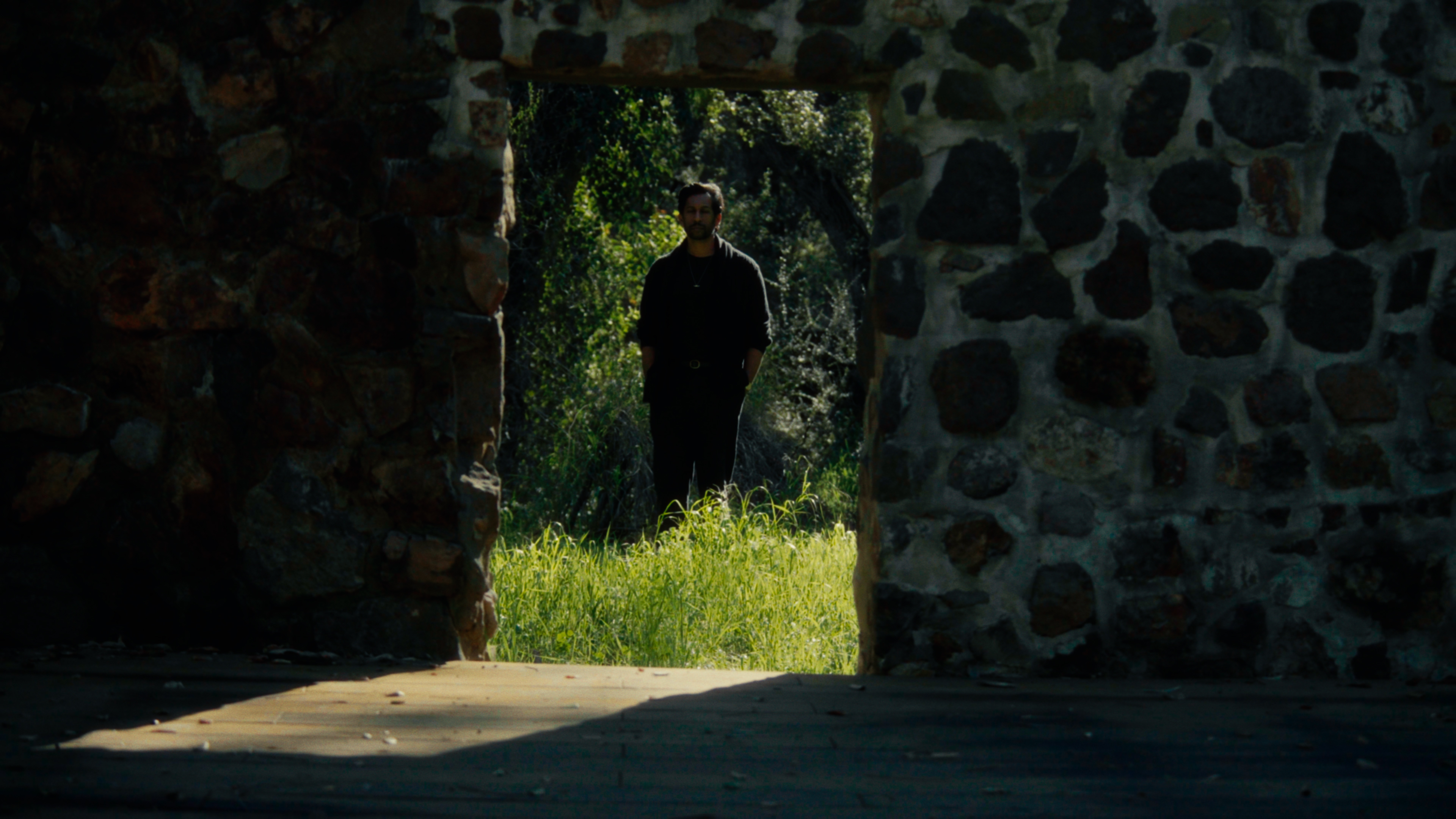 Man standing in a sunlit doorway framed by a stone structure and surrounding greenery