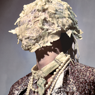 Model wearing sculptural cream headpiece, choker, and beads with a floral lace garment.