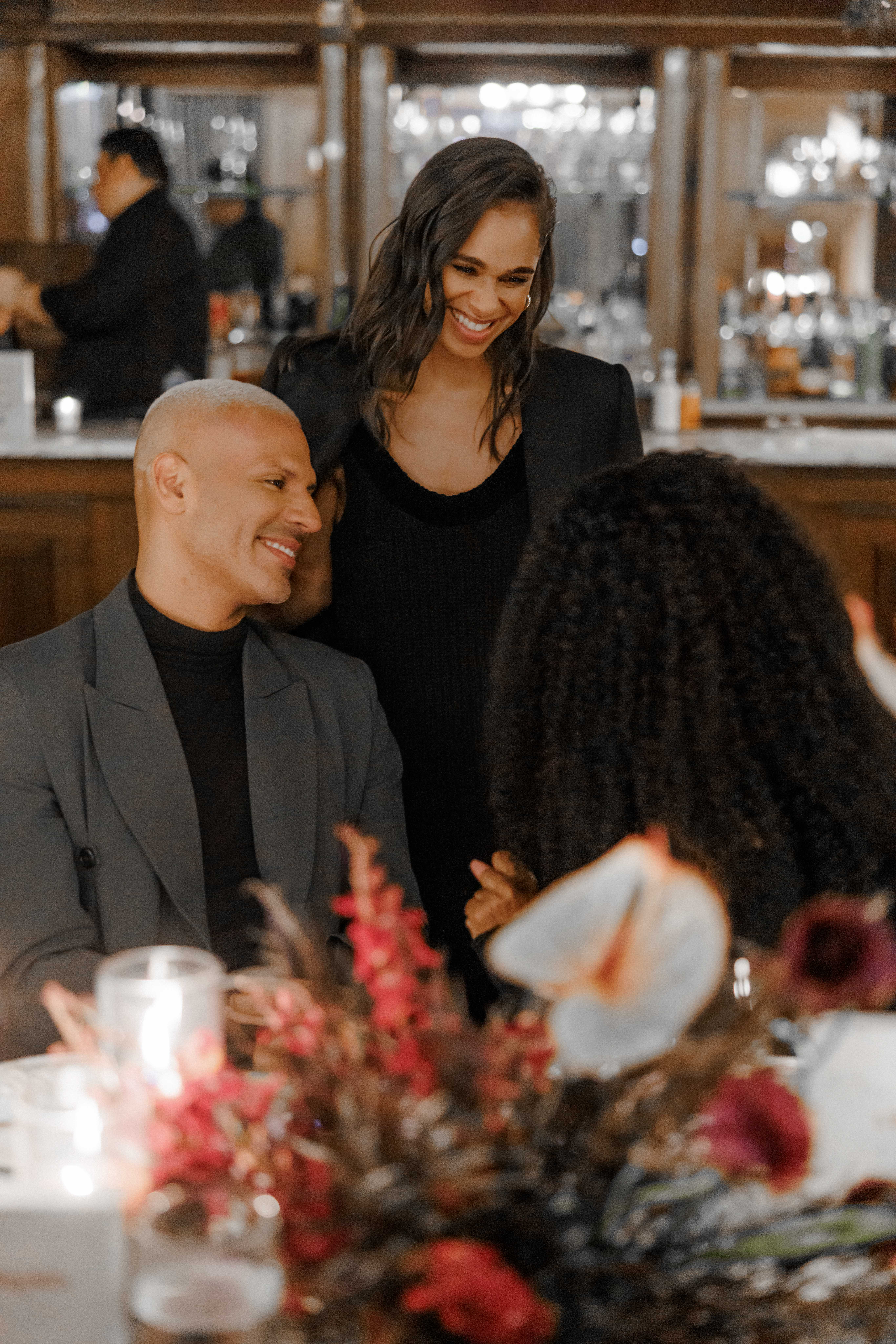 - Three people smiling and talking around a dinner table in a warmly lit restaurant