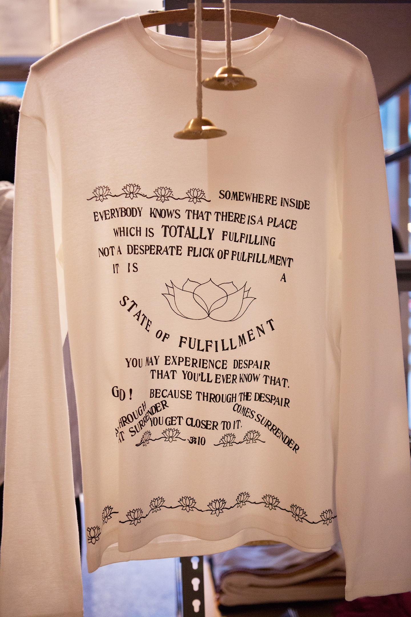White long-sleeve shirt with black text and lotus illustrations hanging on display.