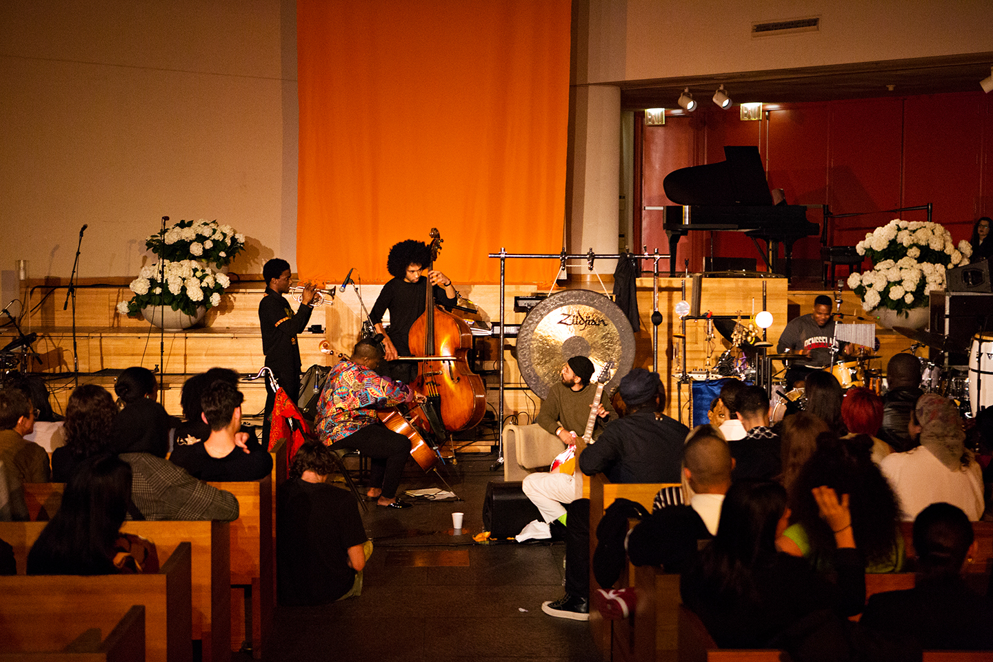 Live music performance in a church-like venue with seated audience and floral stage decor