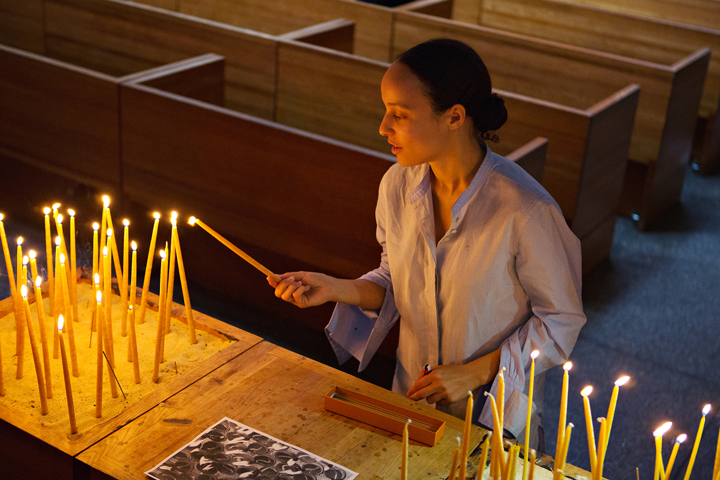  - Person lighting a candle in a church beside rows of glowing candles.