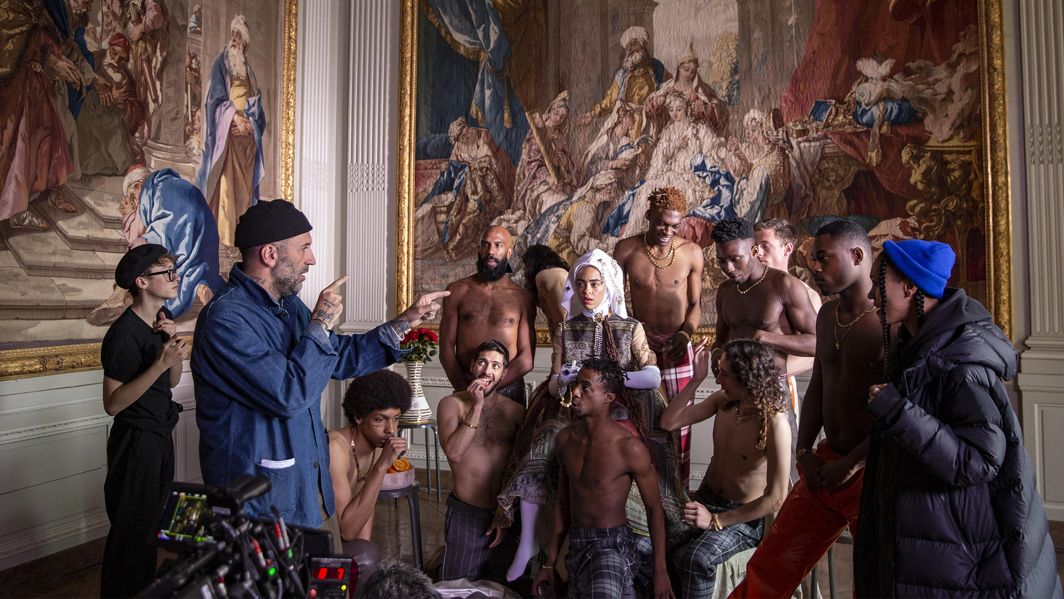 Group posed in ornate room beneath large tapestries, with a director pointing during a shoot.