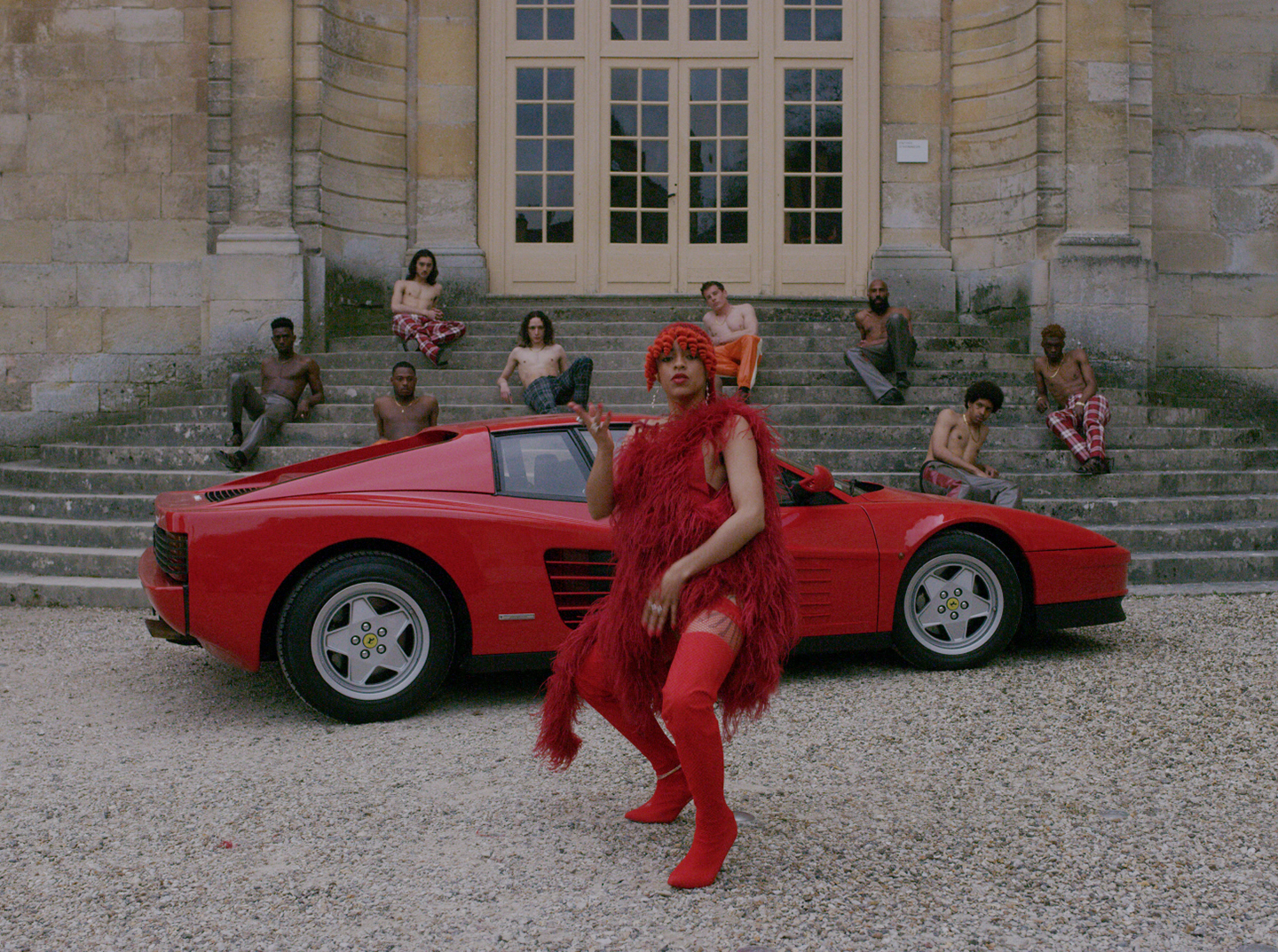 Person in red outfit posing in front of red sports car, with group seated on stone steps.
