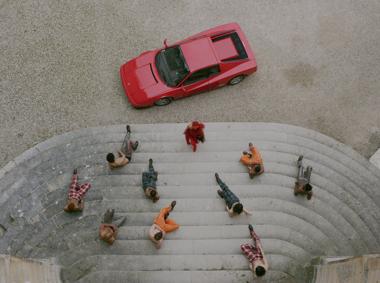  - Overhead view of dancers posed on stone steps beside a red sports car.