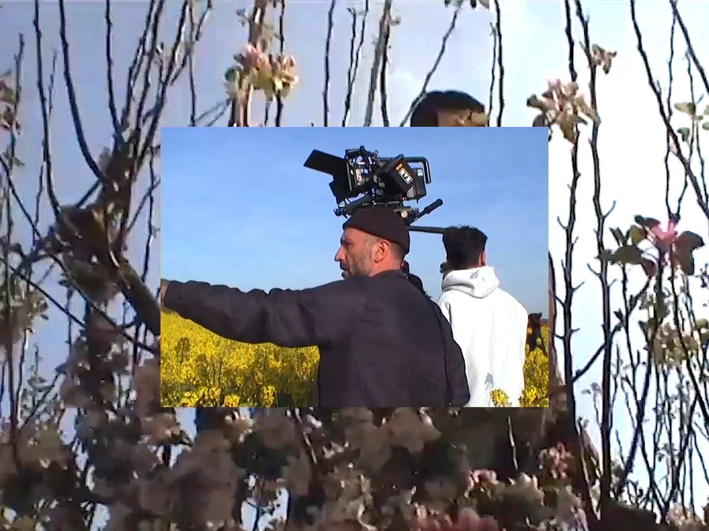 Film crew in a flower field, with camera rig over two people, framed by branches.