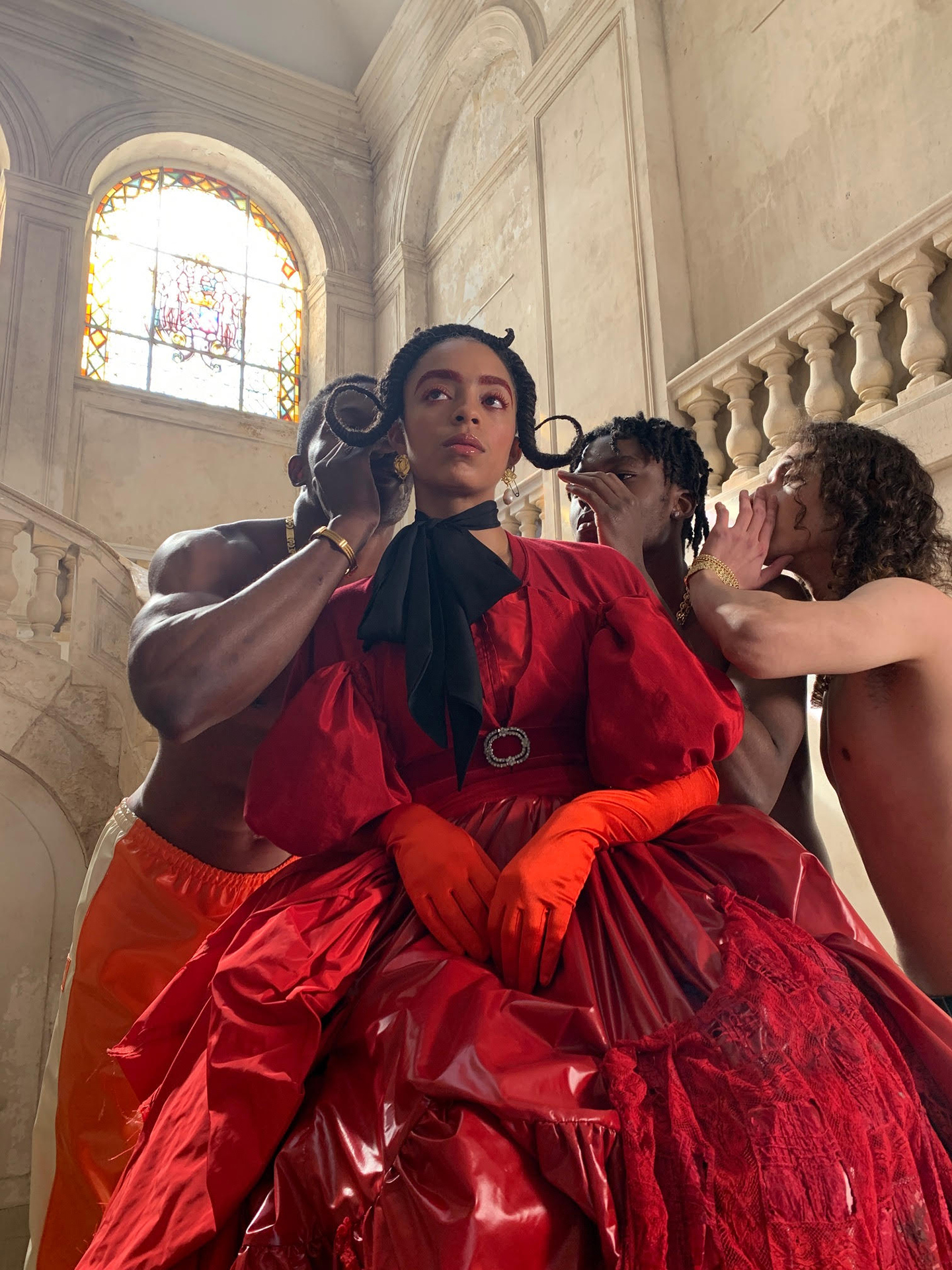 Person in a dramatic red gown posed with three figures in a sunlit stone hall.