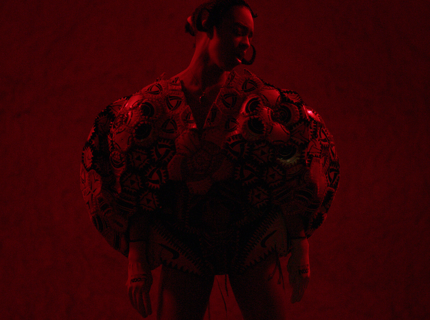 Person in patterned, sculptural outfit under deep red lighting against a textured wall.