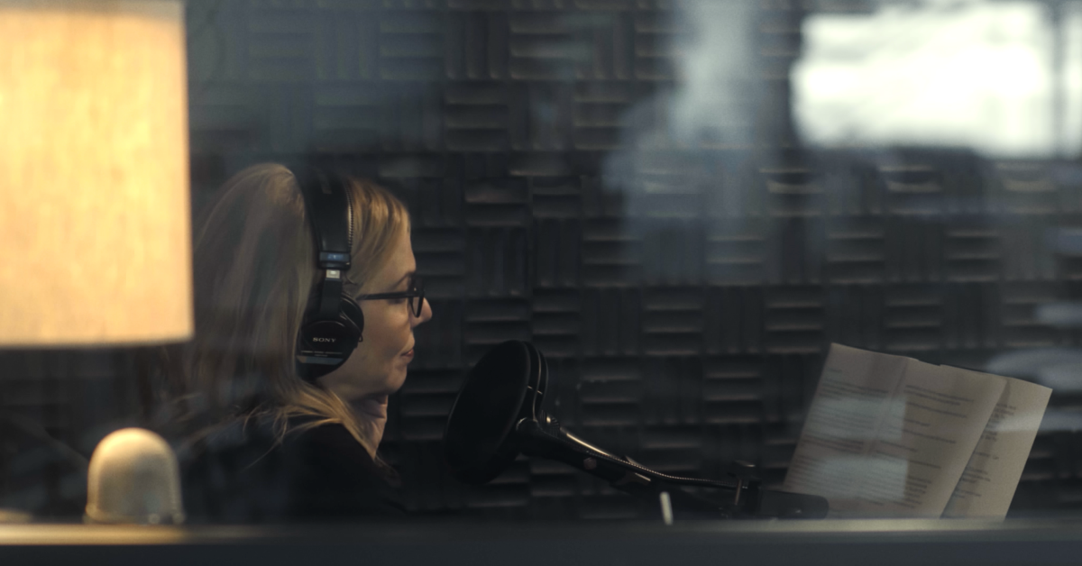 - Person in a recording booth speaking into a microphone, viewed through studio glass.
