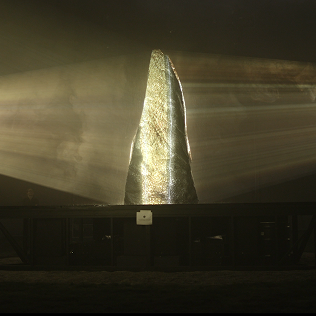 A dark gallery installation with bright light beams passing through a tall sculptural form.