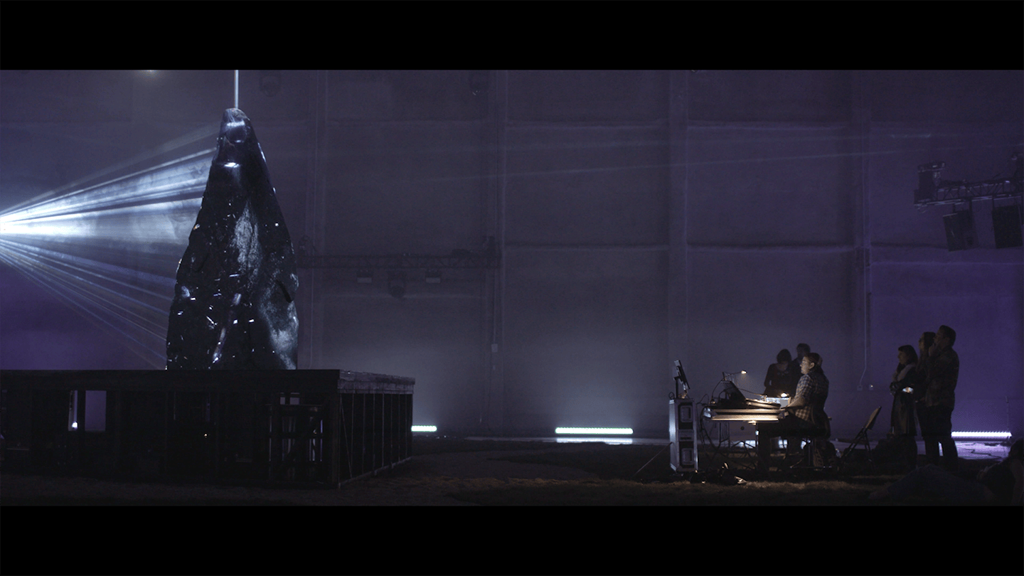 Dark stone monolith lit by projector in a dim warehouse as crew watches.