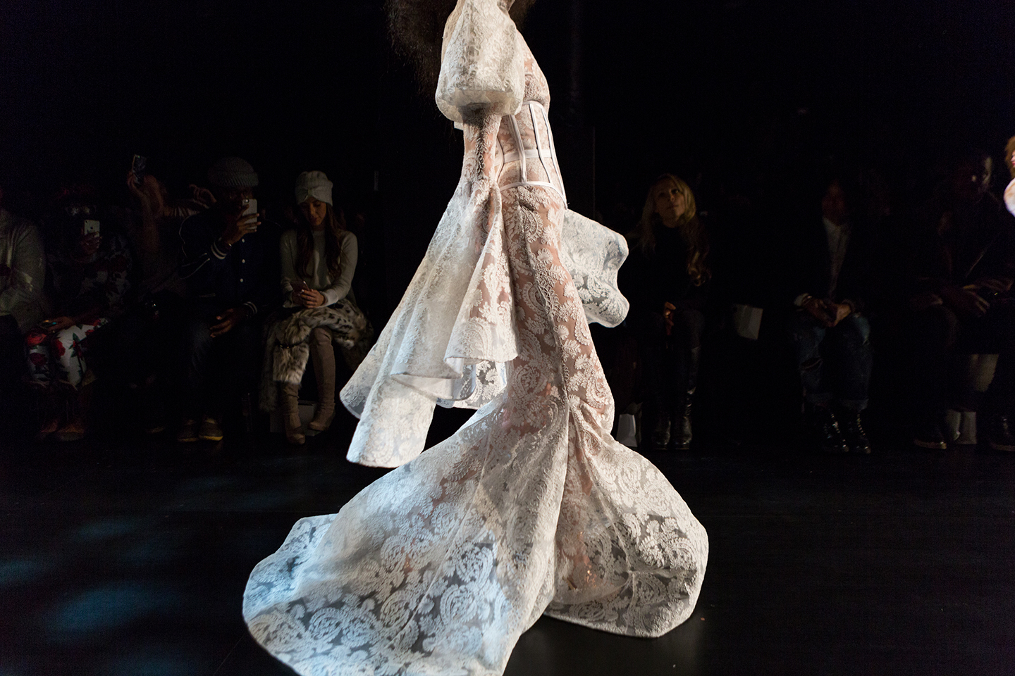 Model in flowing white lace gown walking a dark runway