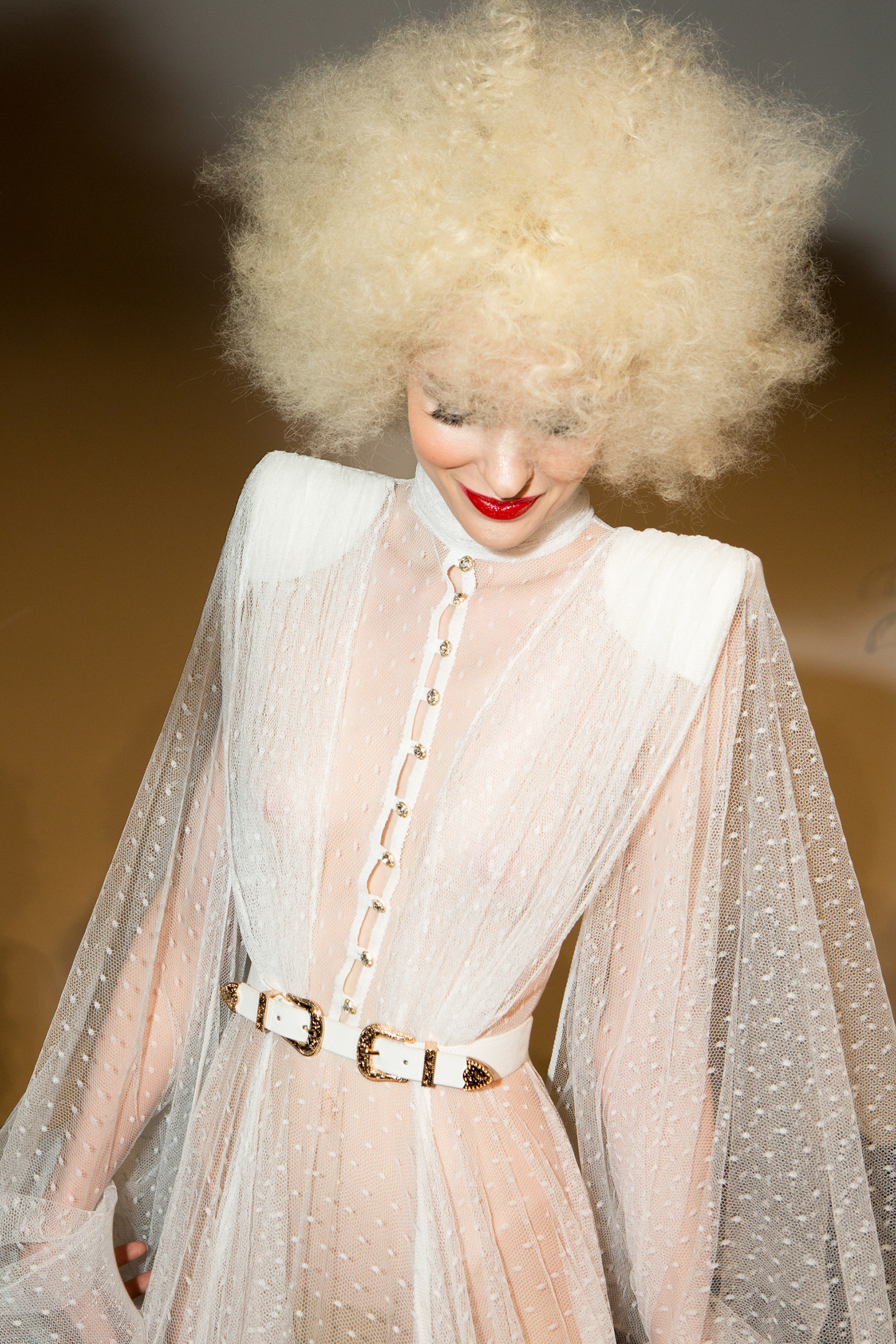 Model in sheer blush gown with white fringe overlay and belt.