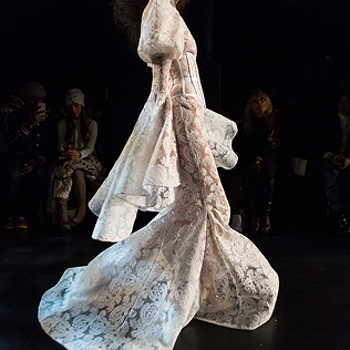 Model in flowing white lace gown walking a dark runway.