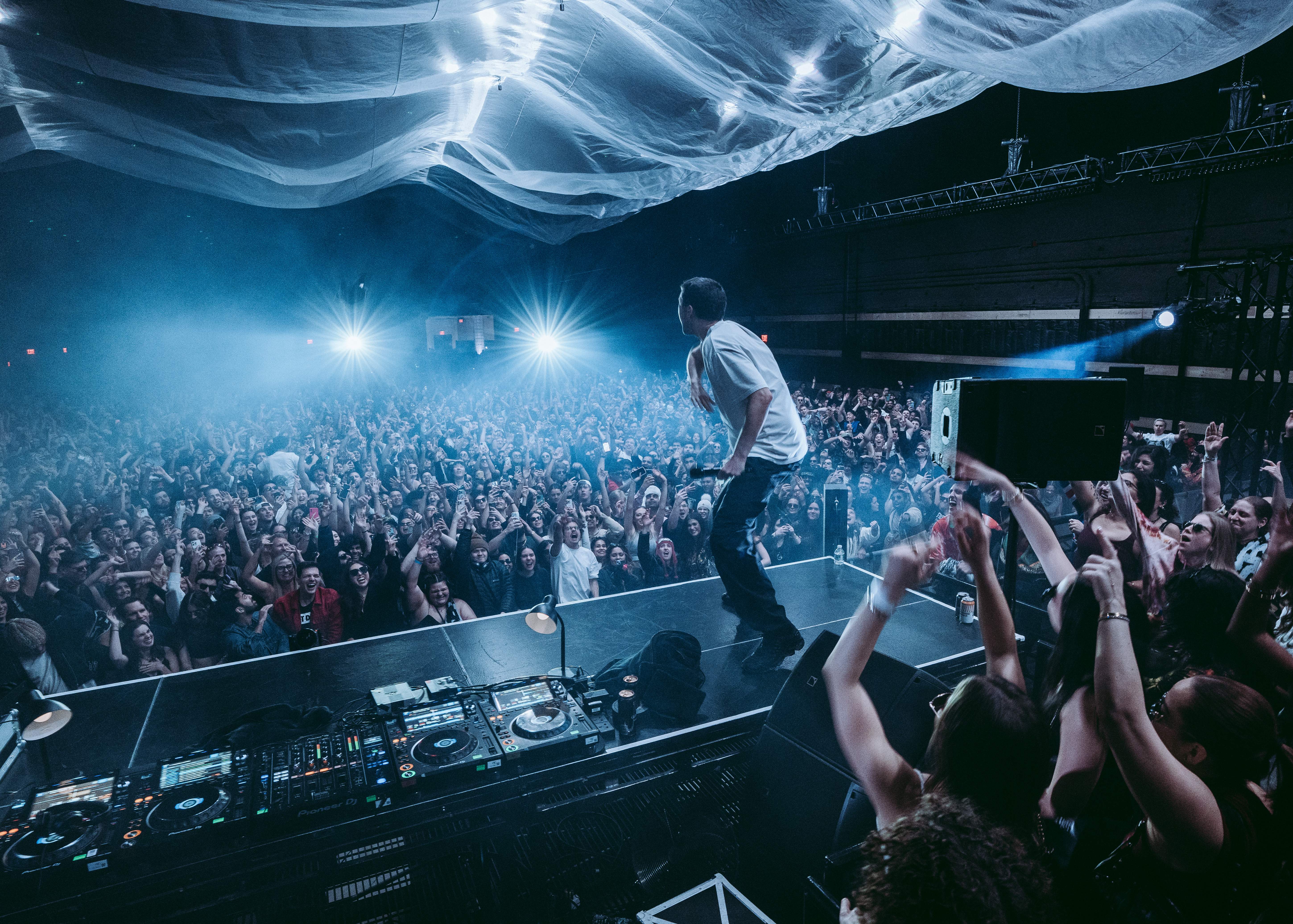 DJ performing on stage in front of a large crowd beneath flowing ceiling fabric and blue lights