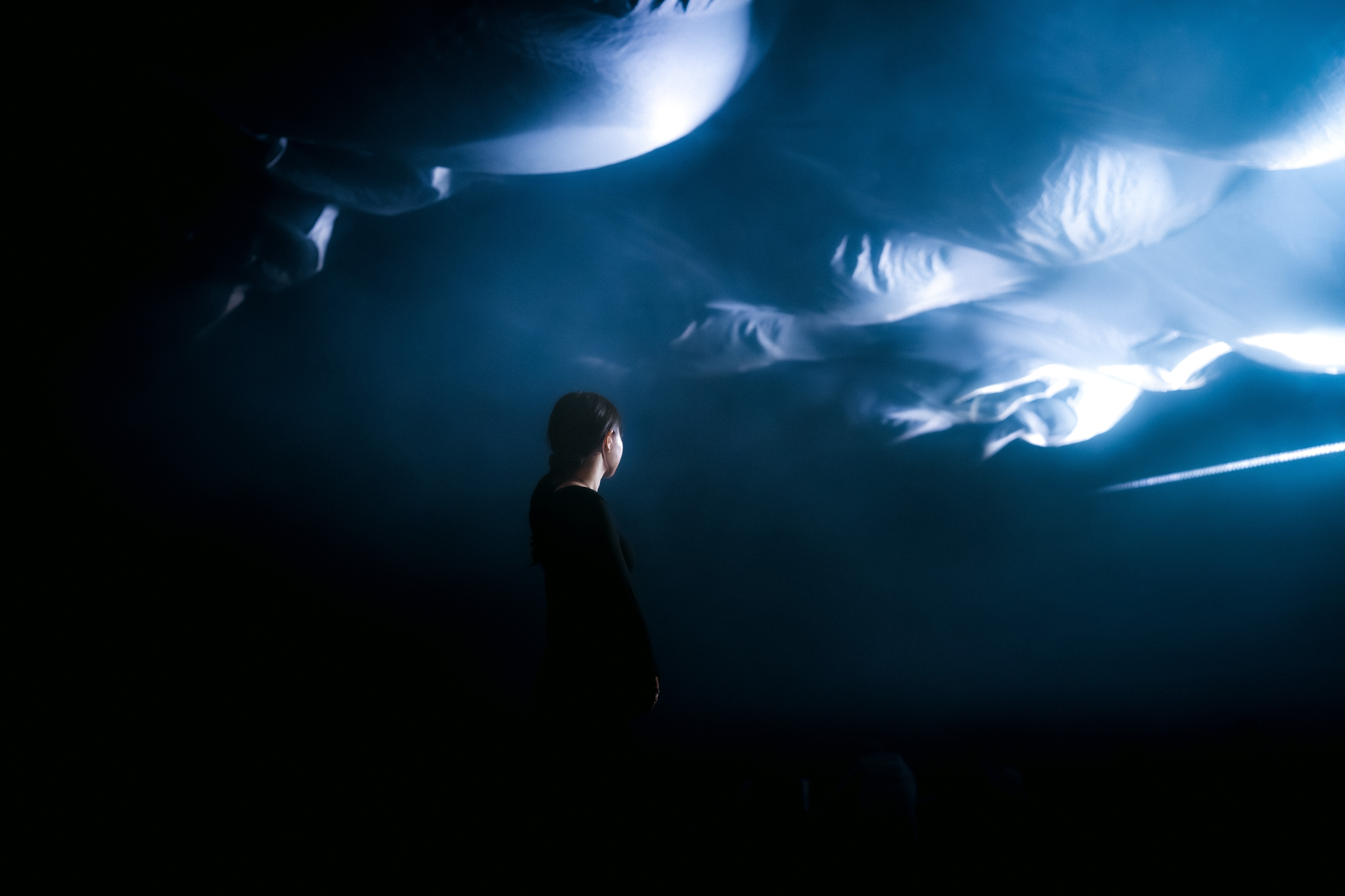 Person standing beneath glowing blue fabric installation in a dark indoor space