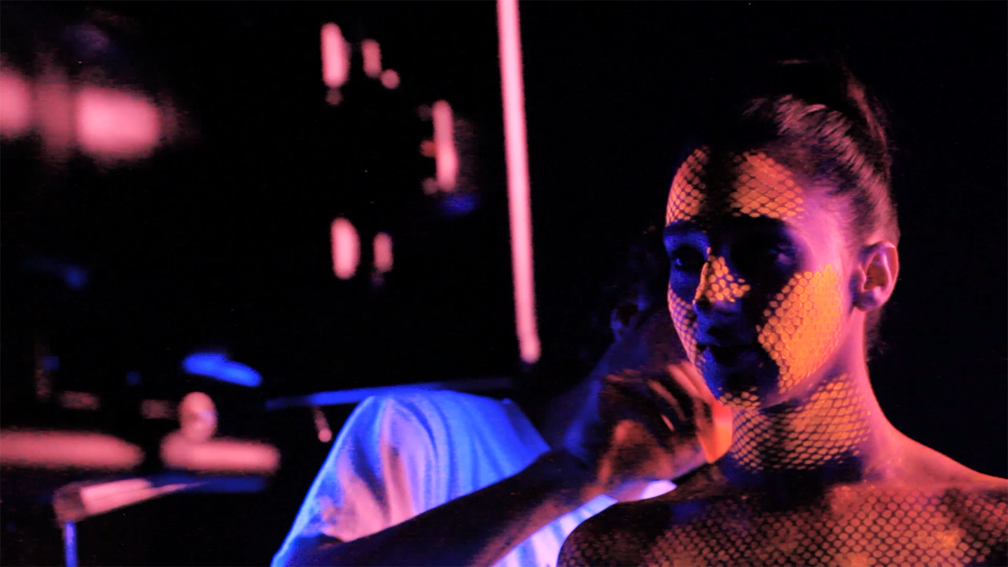 A person lit in pink, orange, and blue stands against a dark background, with a honeycomb-like pattern of light across their face and body while another person is partially visible behind them.