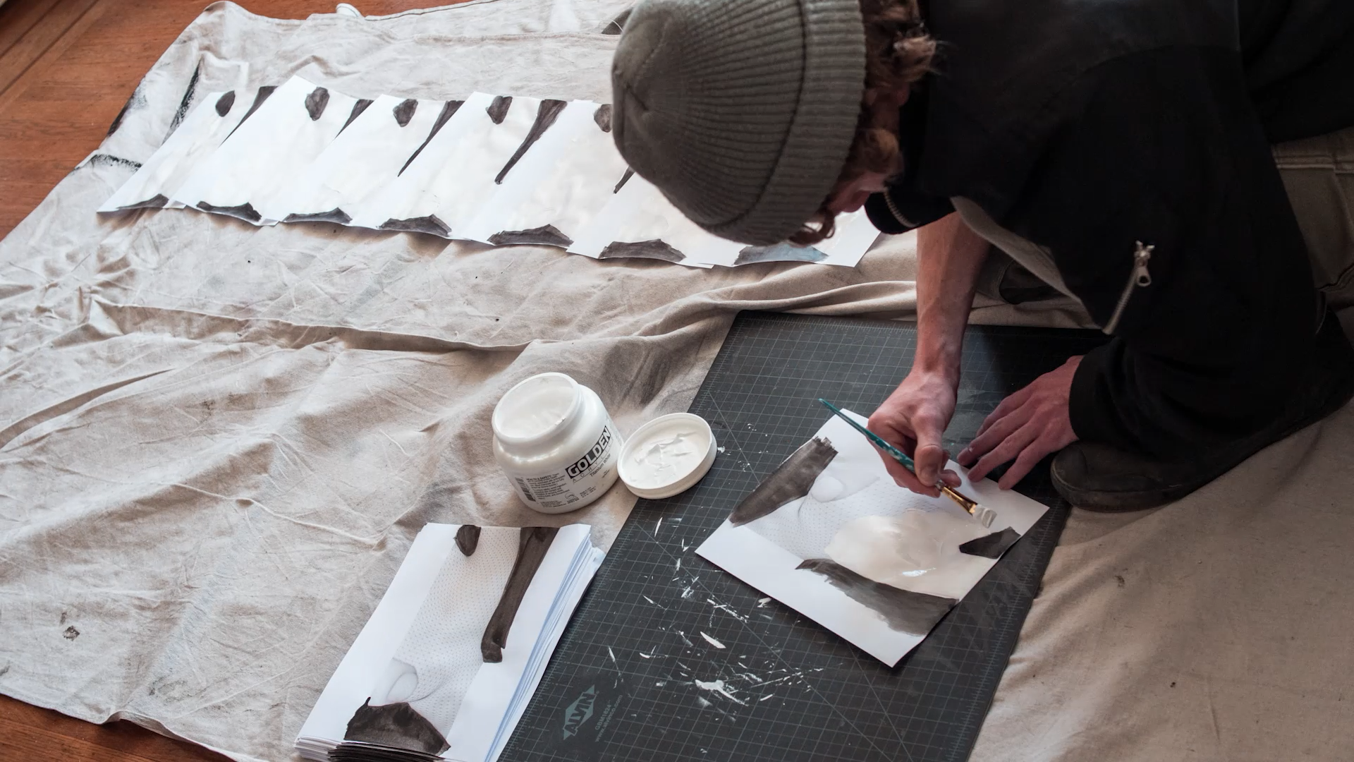 A person in a beanie paints black and white shapes on paper laid out over a drop cloth, with a jar of white paint and stacked papers nearby.