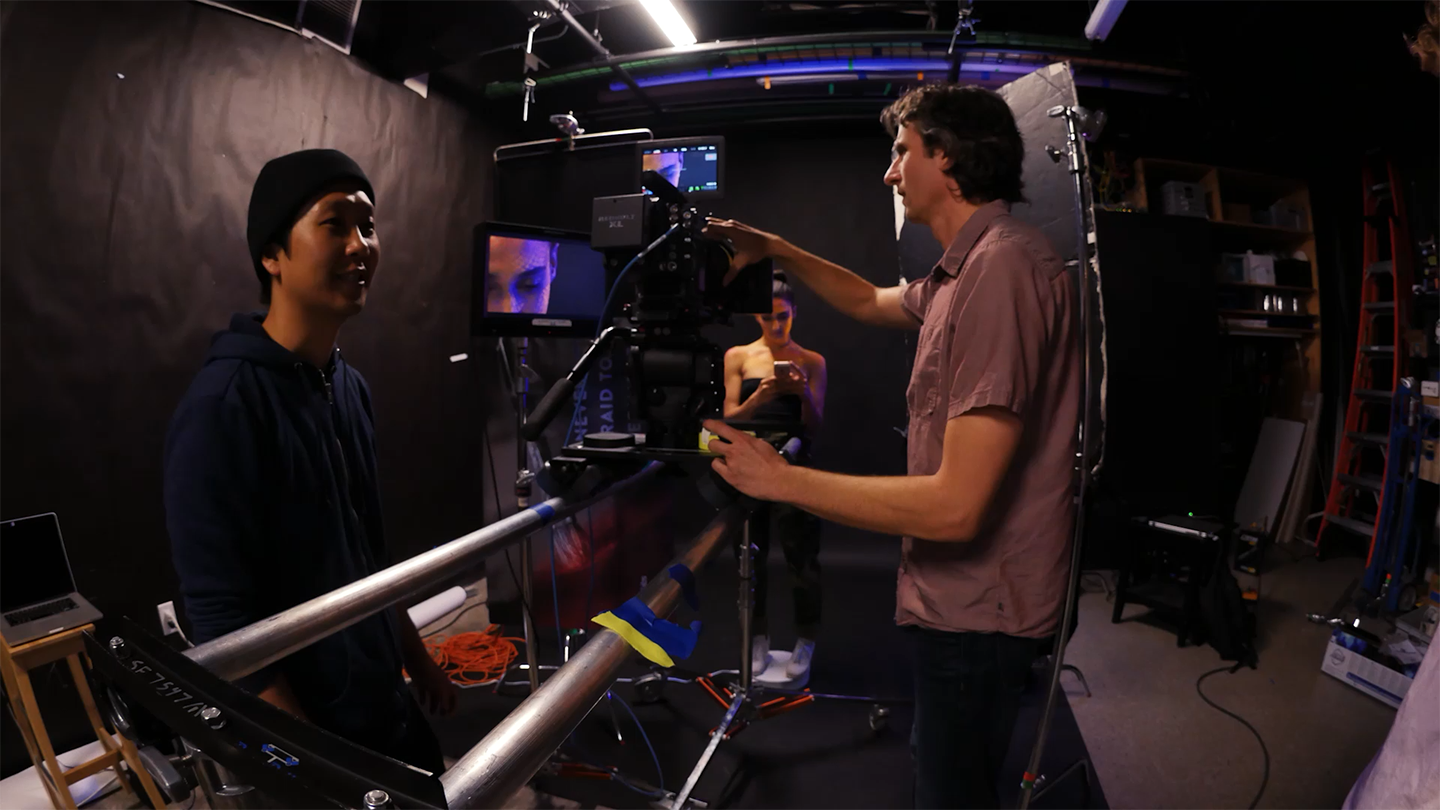 A film crew is setting up a camera on a studio rig, with one person adjusting the equipment, another standing nearby, and a third person visible in the background behind the camera.