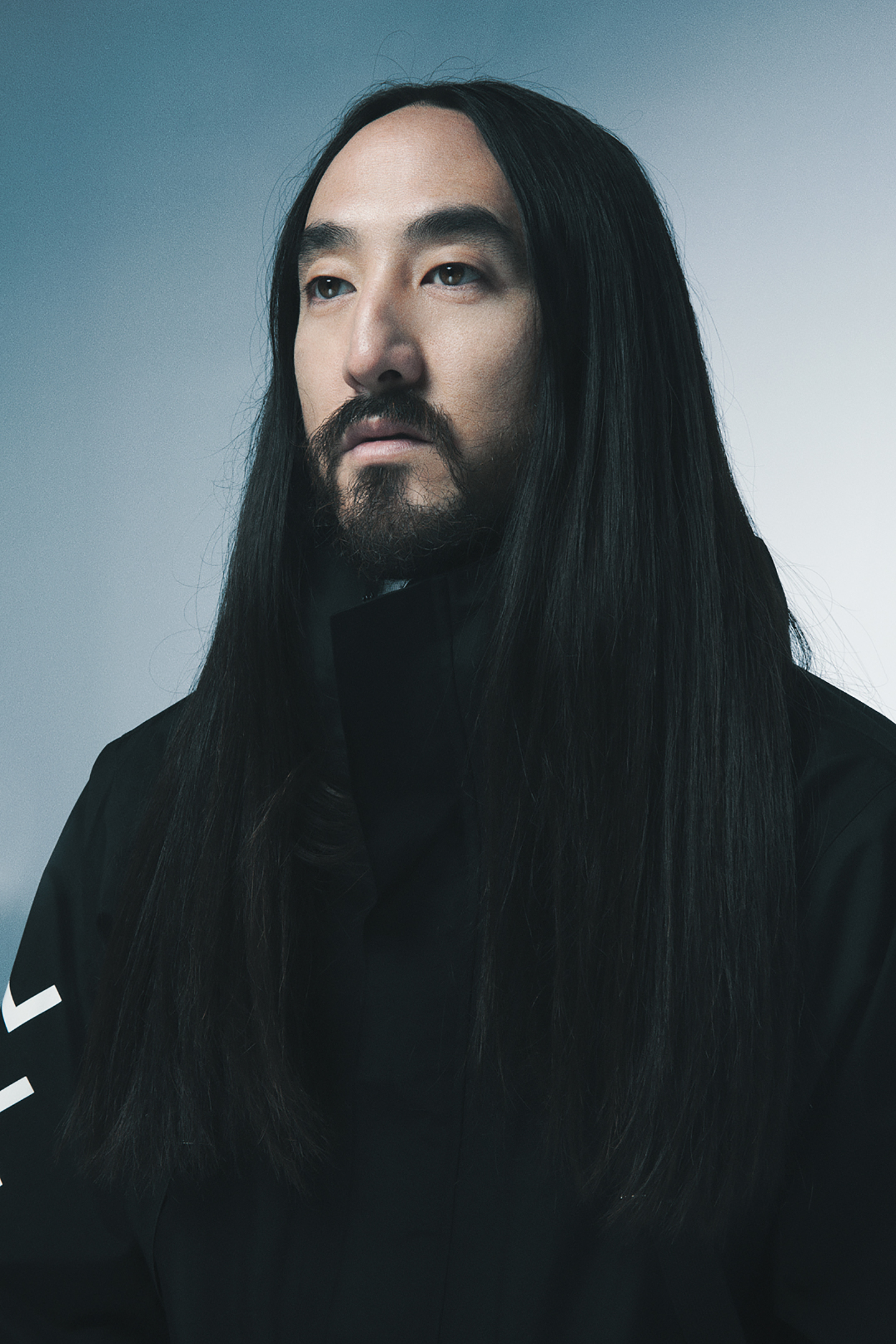 Portrait of a person with long black hair and a beard, wearing a dark jacket against a gray background.