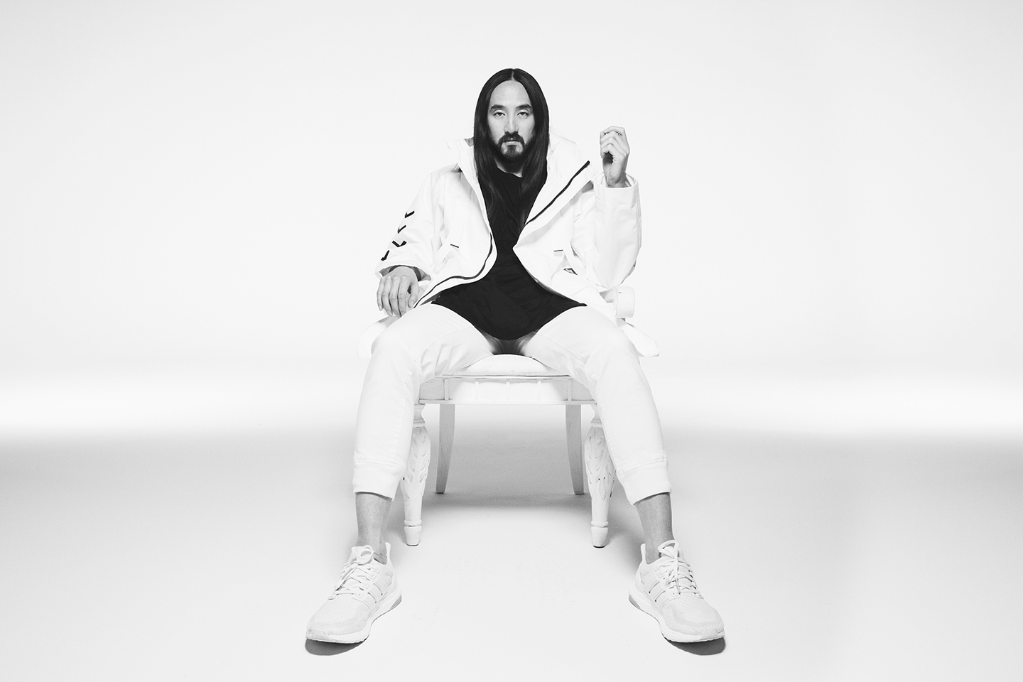 A black-and-white studio portrait of a man with long dark hair sitting on a white chair against a plain white background, wearing a white jacket and pants over a dark shirt and white sneakers.