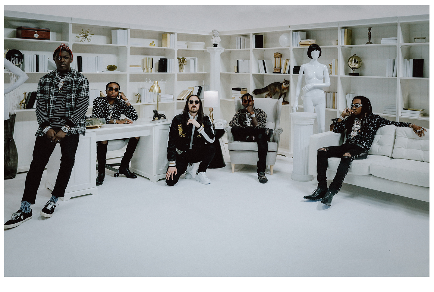 Five men pose in a bright white studio-like room with built-in shelves and decorative objects, one standing and the others seated or crouching in dark patterned outfits and sunglasses.
