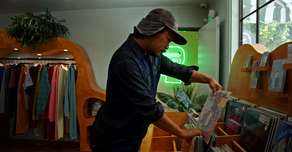 A man in a wide-brimmed hat browses vinyl records in a warmly lit shop, with clothing racks, plants, and a neon sign in the background.