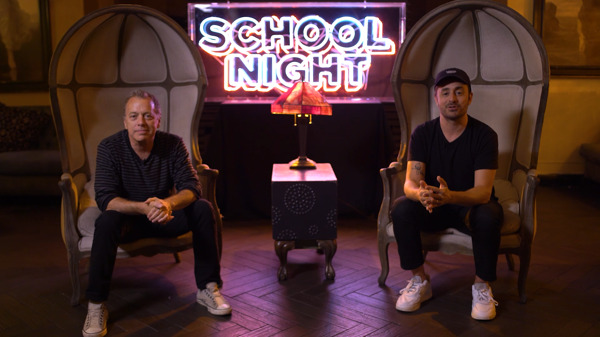 Two men sit in oversized chairs on a stage with a neon “SCHOOL NIGHT” sign glowing behind them and a small lamp on a table between the chairs.