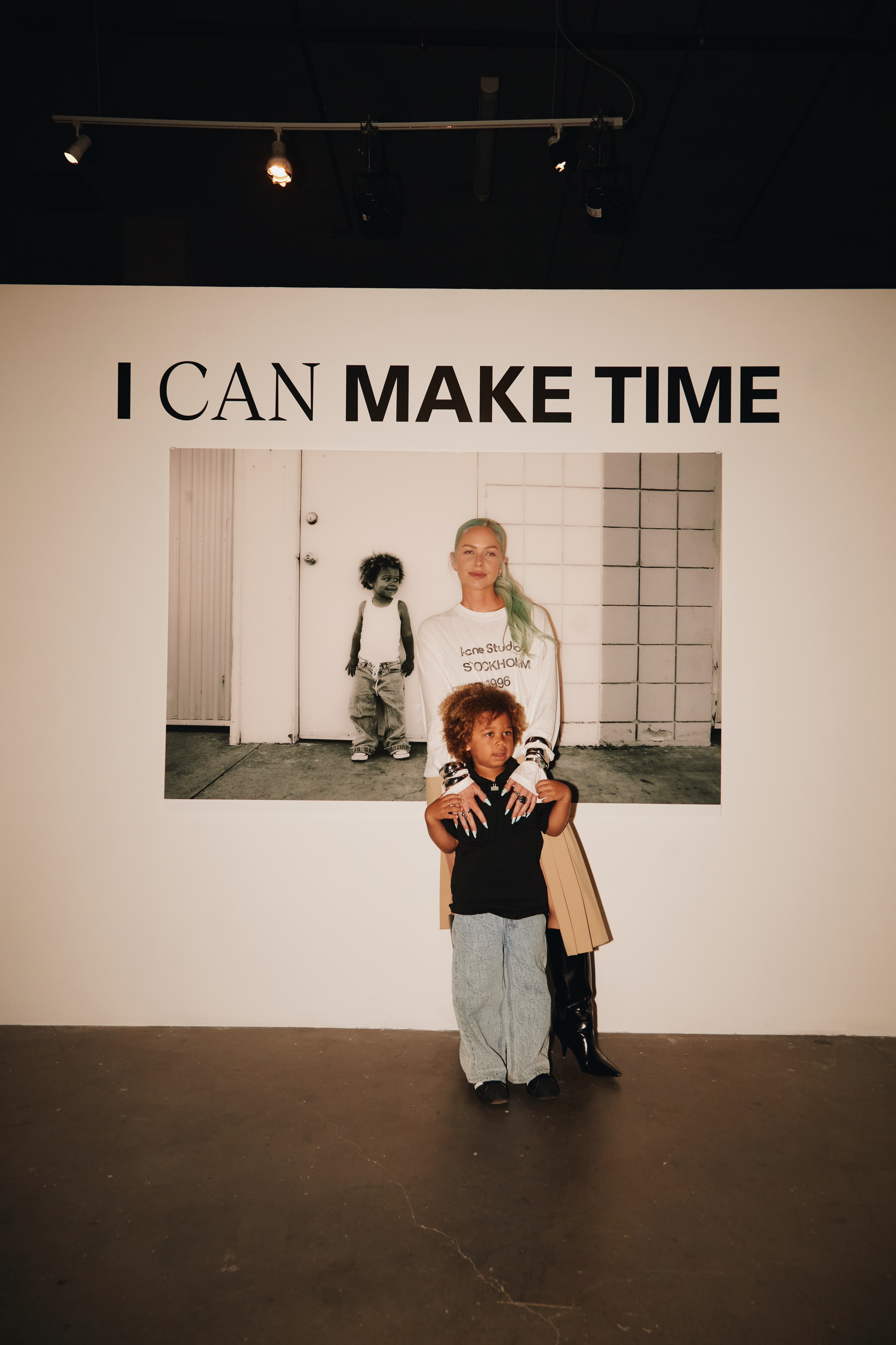 Photographer poses with child beneath I Can Make Time title and large portrait at exhibition