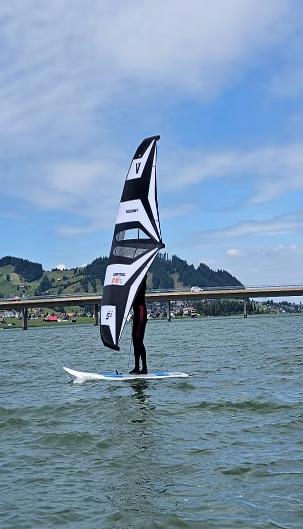 Surfer auf einem See mit Bergen und einer Brücke im Hintergrund