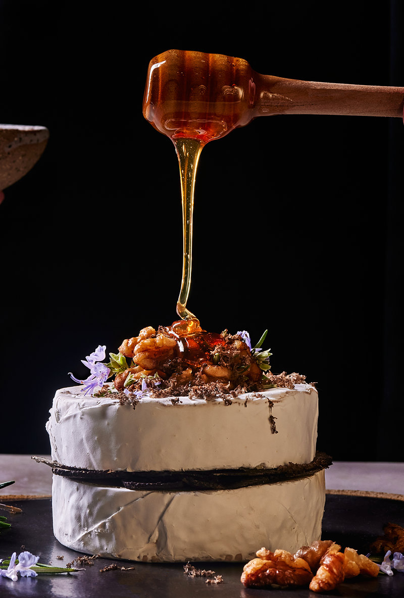 Honey being drizzled from a wooden dipper onto a wheel of Brie cheese topped with walnuts and truffles.