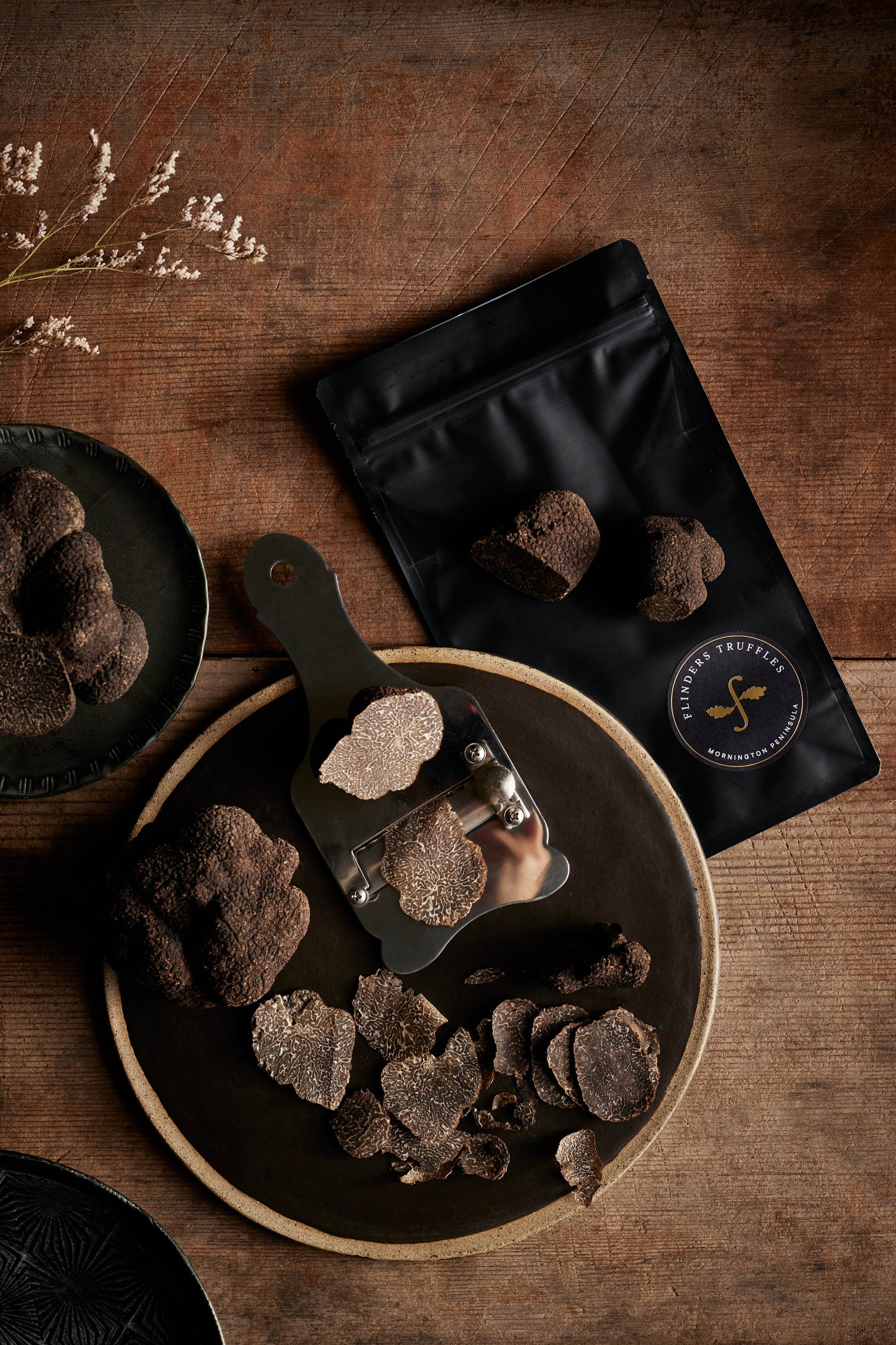 Sliced black truffles on a black plate with a truffle slicer and packaged truffles on a wooden table.