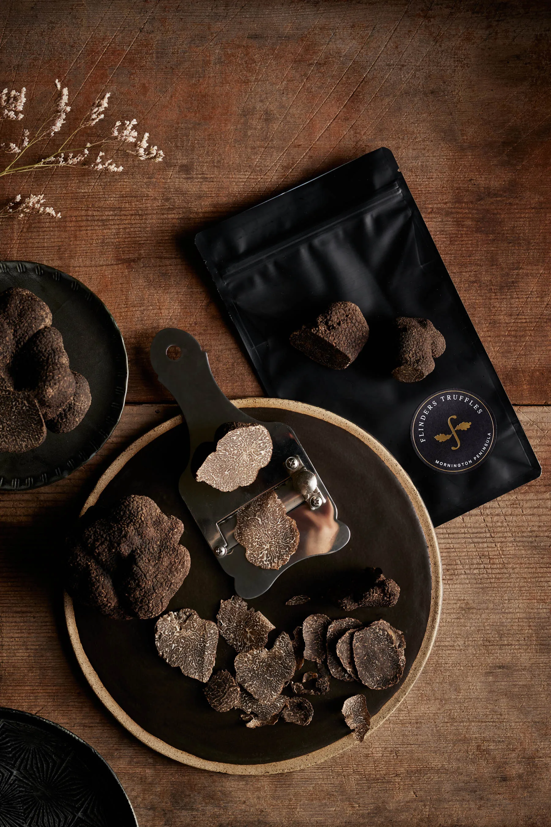 Sliced black truffles on a black plate with a truffle slicer and packaged truffles on a wooden table.