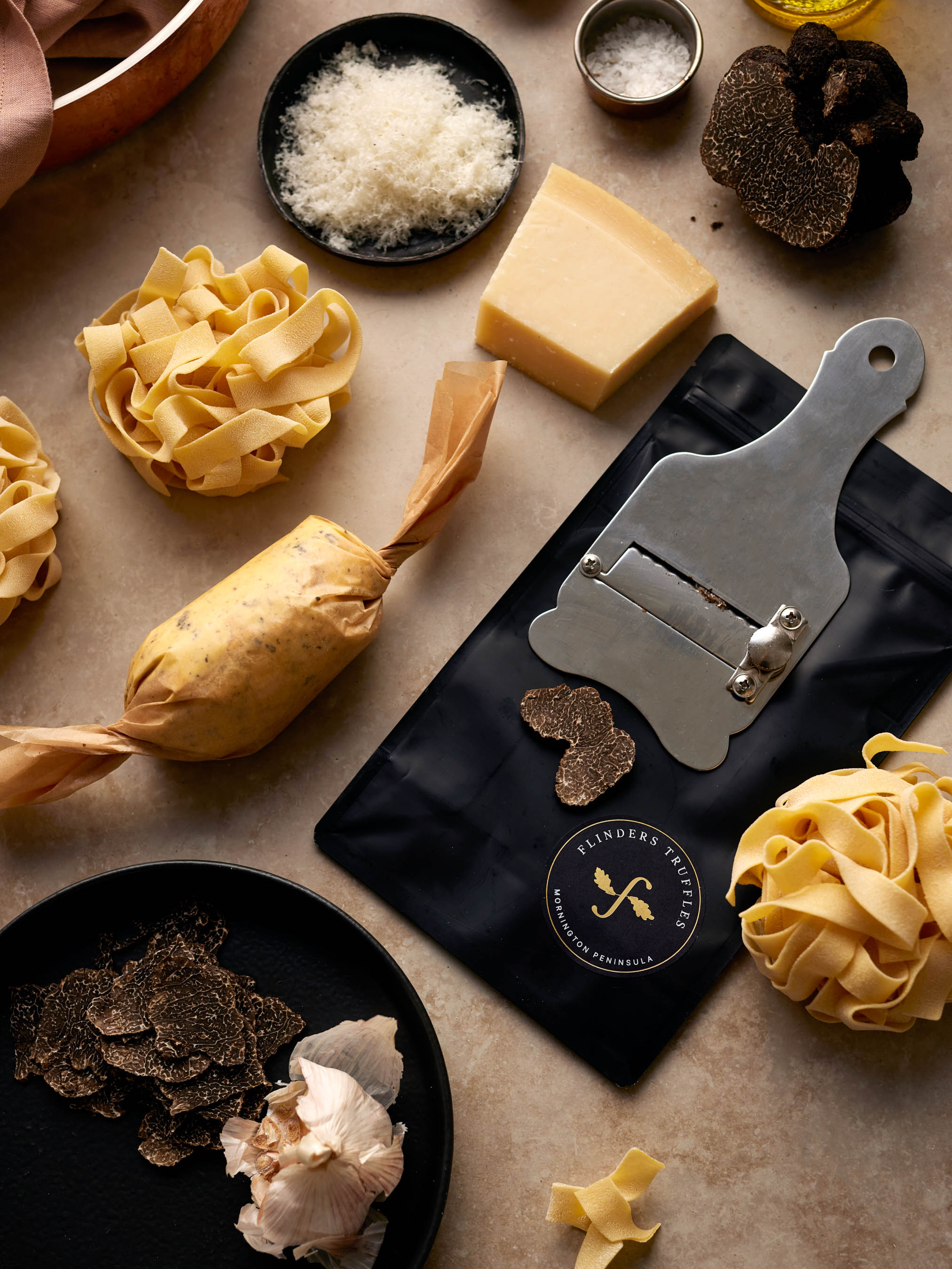 Flat lay of fresh tagliatelle pasta, black truffles, cheese, garlic, truffle slicer, and truffle butter on a beige surface.