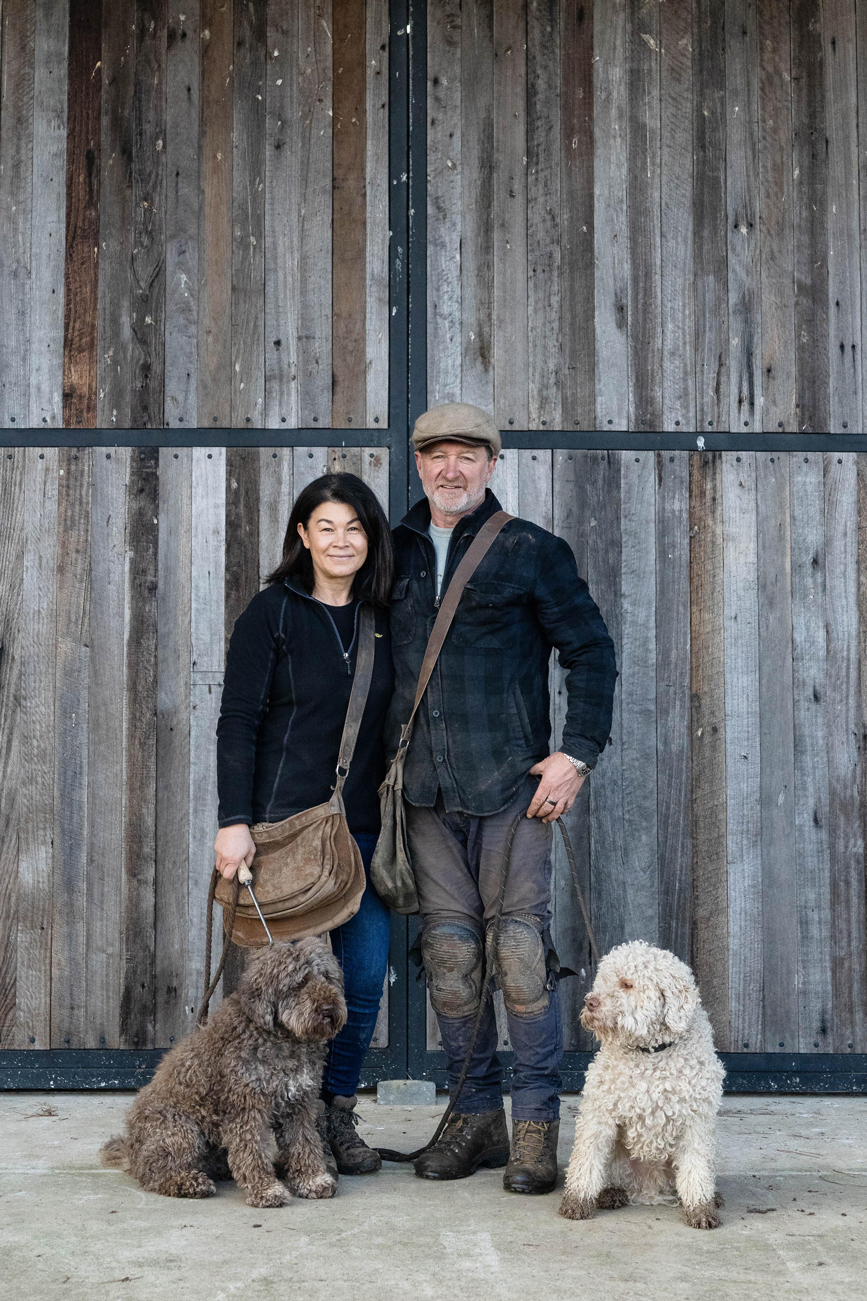 Man and woman standing in front of a wooden gate with two curly-haired dogs sitting beside them.
